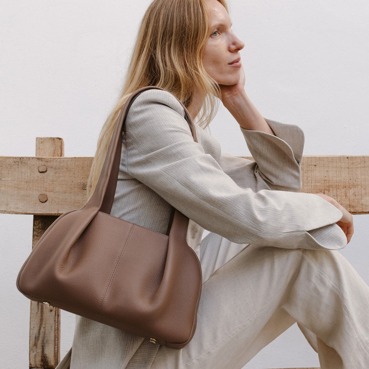 Model sat on a bench with STOW London's Bowling bag in Mocha pebbled leather featuring the pleated front panel and gold-coated stainless steel hardware