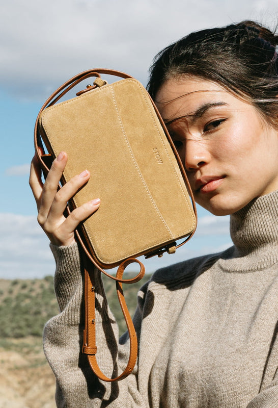 STOW Soft Sand Suede Camera Bag being held by model against green hills and blue skies.