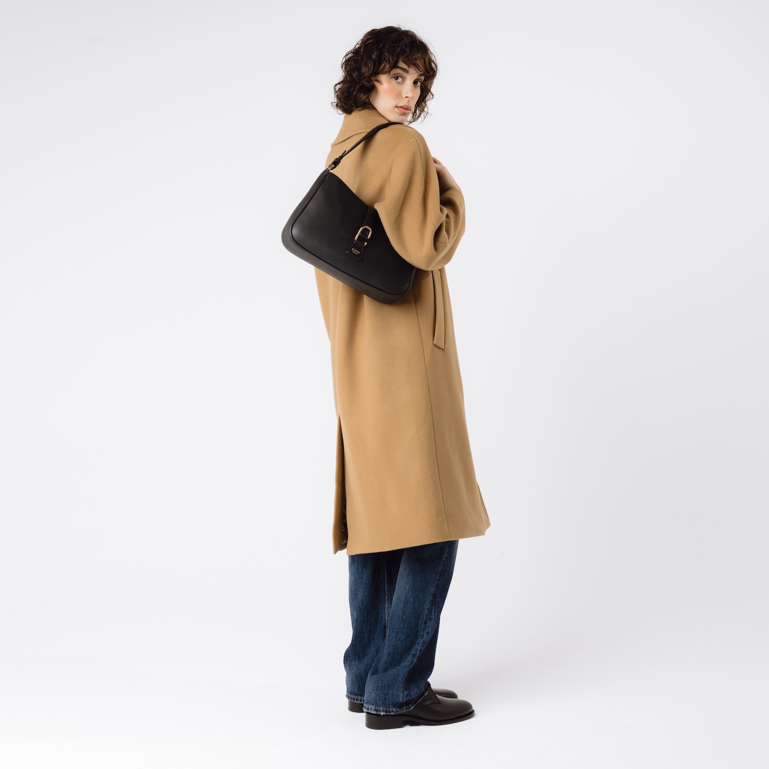 Model wearing STOW London's Beth bag in Black pebbled and smooth leather on her shoulder featuring gold-coated stainless steel hardware and clip on a white background