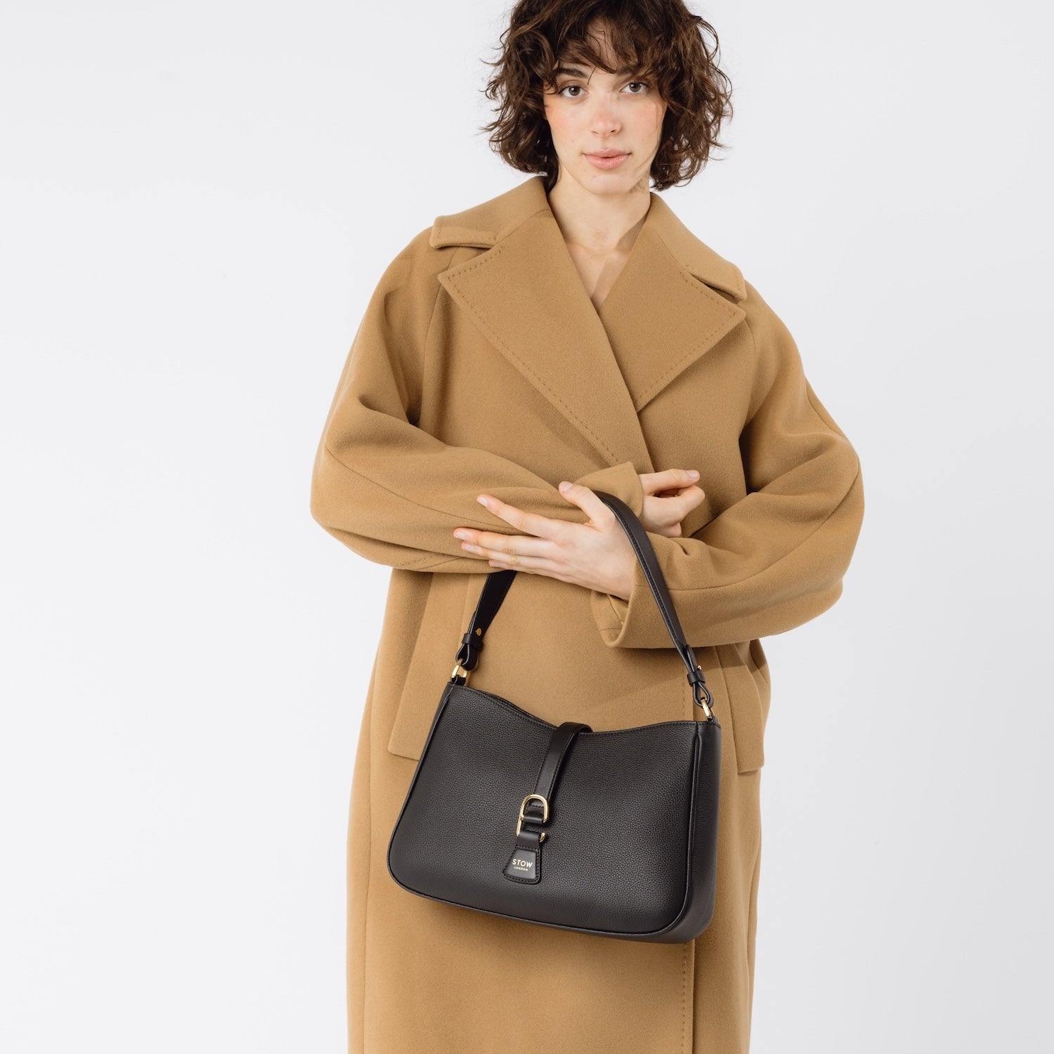 Model holding STOW London's Beth bag in Black pebbled and smooth leather in her arms at her front featuring gold-coated stainless steel hardware and clip on a white background