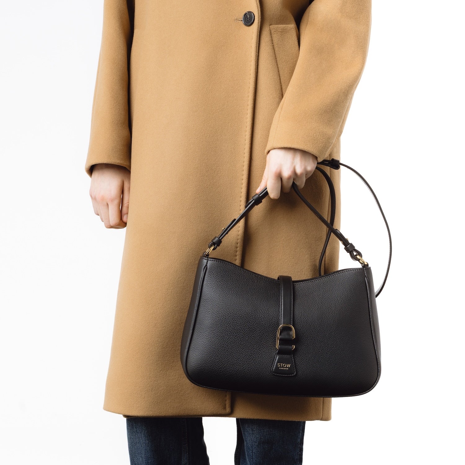 Model holding STOW London's Beth bag and detachable crossbody strap in Black leather in her hand at her front featuring gold stainless steel hardware on a white background