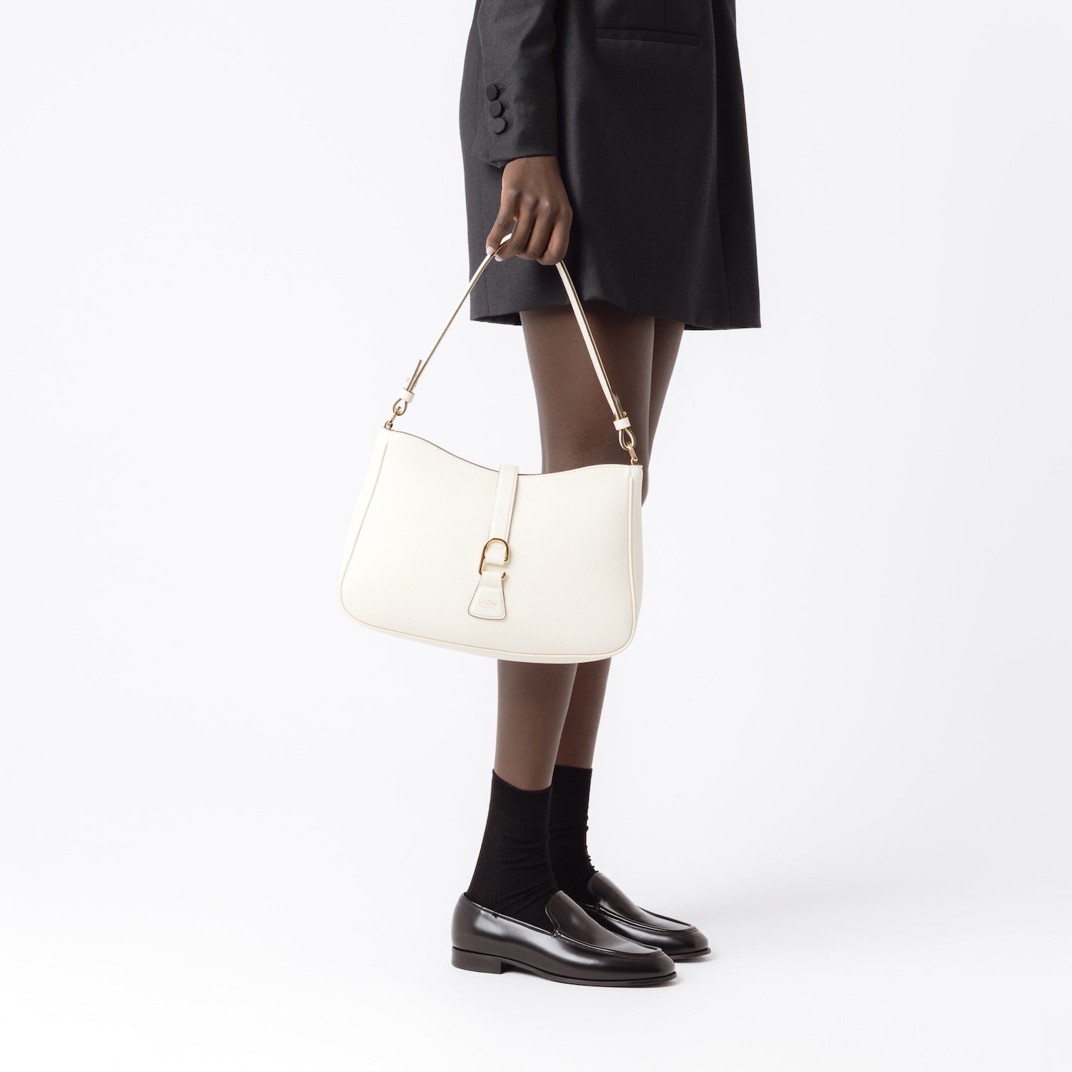 Model holding STOW London's Beth bag in Spring Moon pebbled and smooth leather in her hand at her side featuring gold-coated stainless steel hardware and clip on a white background