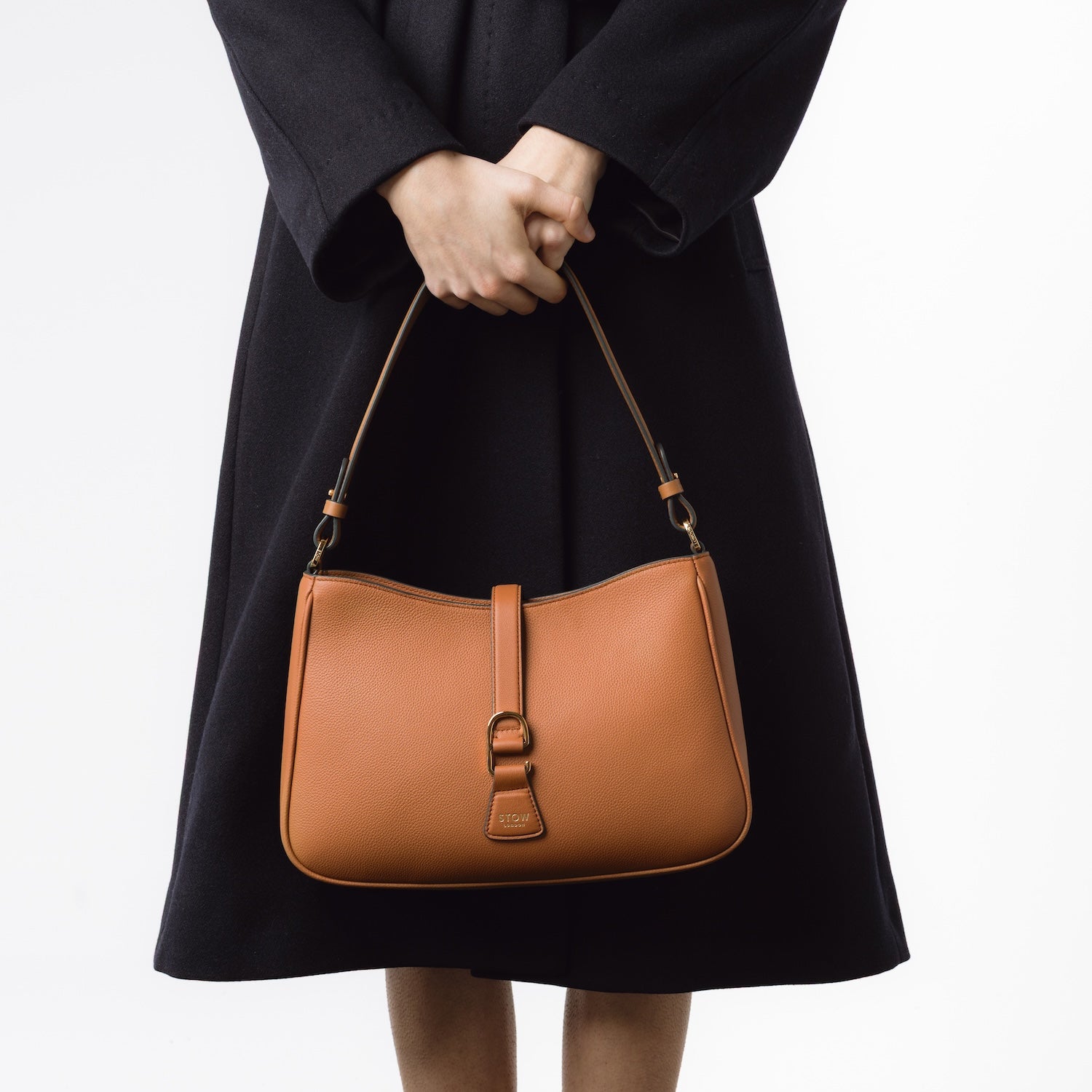 Model holding STOW London's Beth bag in Earth Tan pebbled leather at her front featuring gold-coated stainless steel hardware on a white background