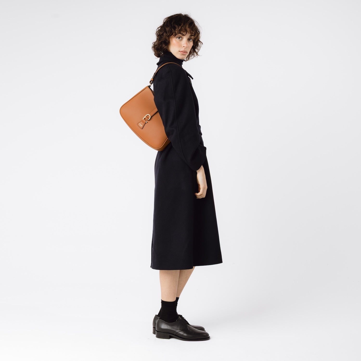 Model wearing STOW London's Beth bag in Earth Tan pebbled leather on her shoulder featuring gold-coated stainless steel hardware on a white background