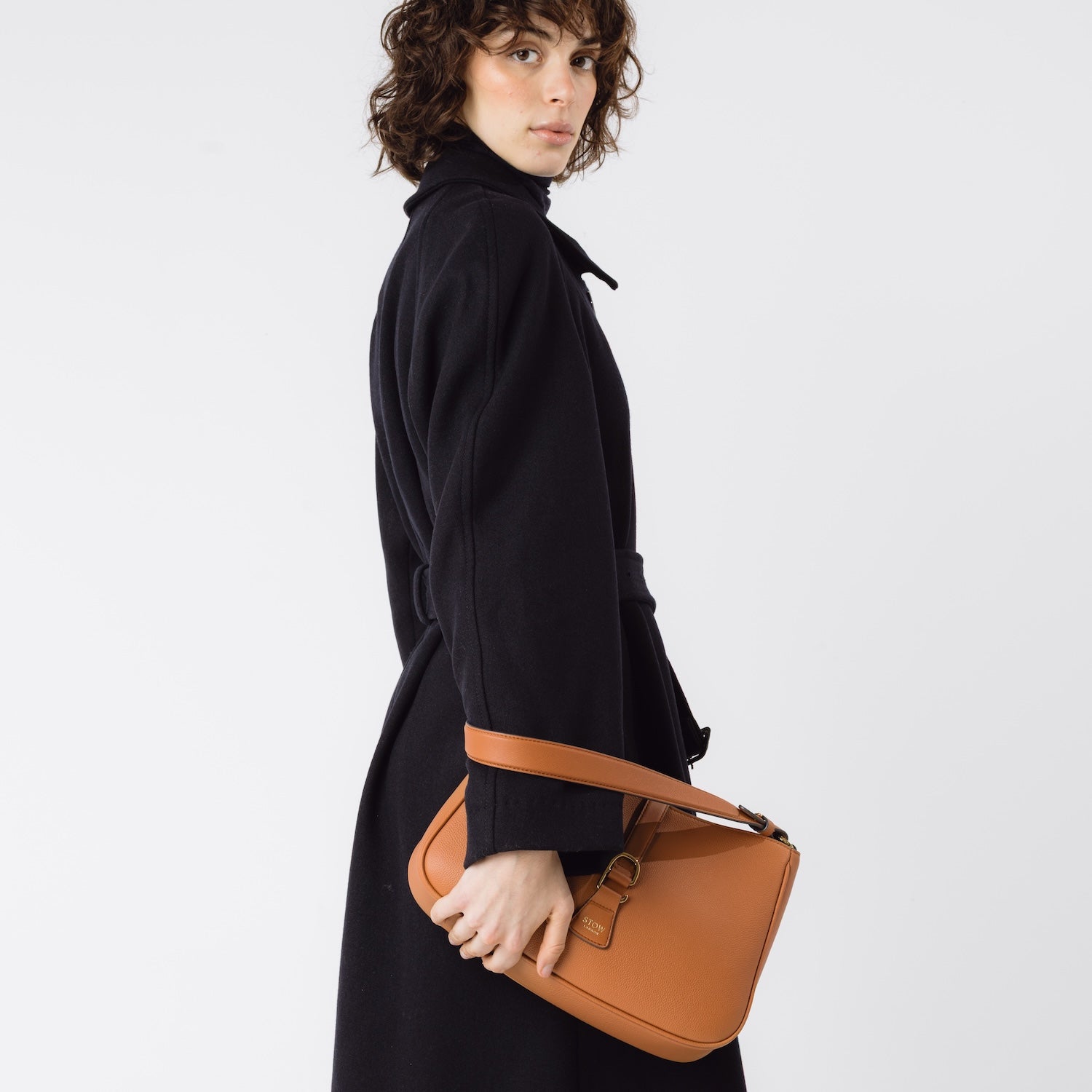 Model holding STOW London's Beth bag in Earth Tan pebbled and smooth leather in her hand at her side featuring gold-coated stainless steel hardware on a white background