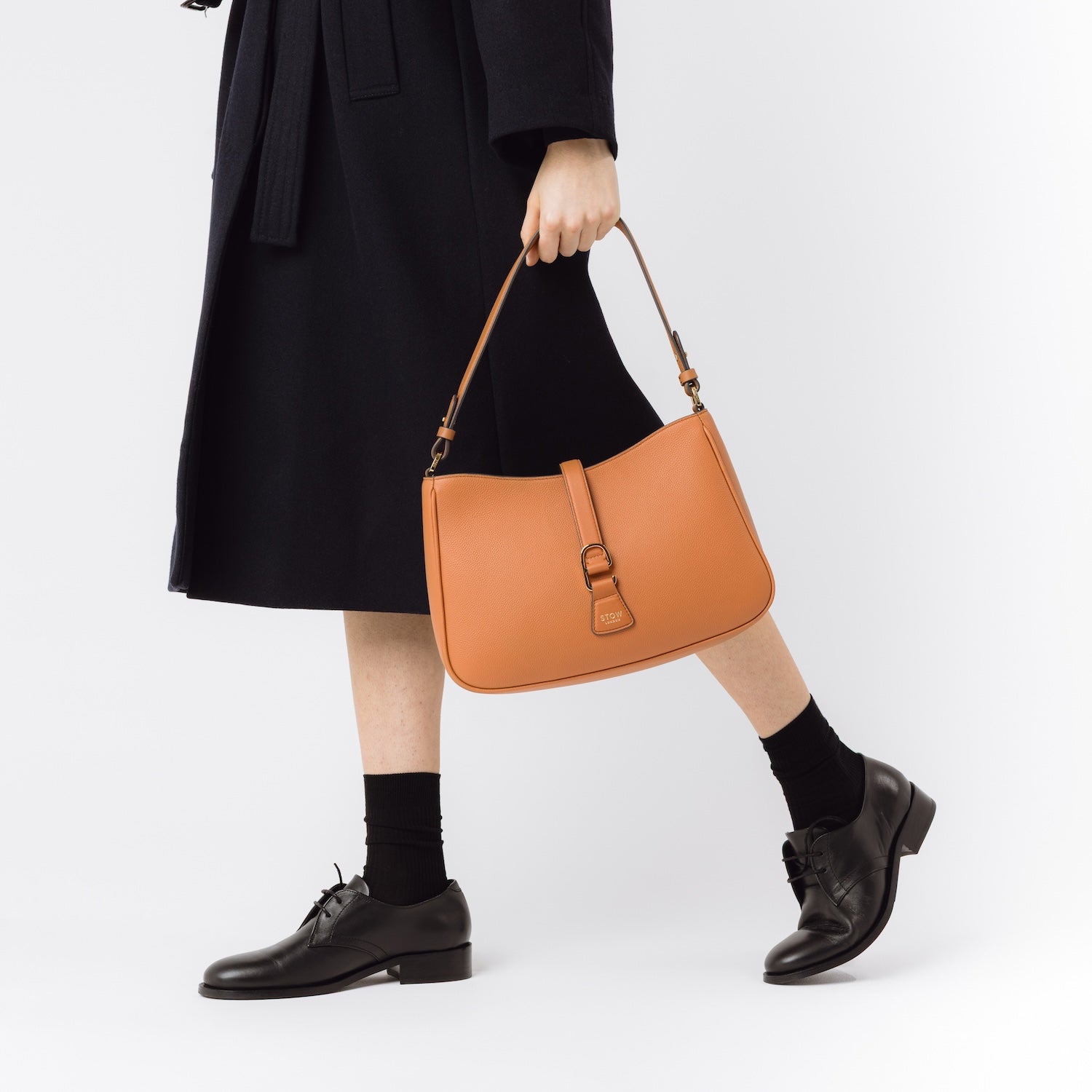 Model holding STOW London's Beth bag in Earth Tan pebbled and smooth leather at her side featuring gold-coated stainless steel hardware on a white background