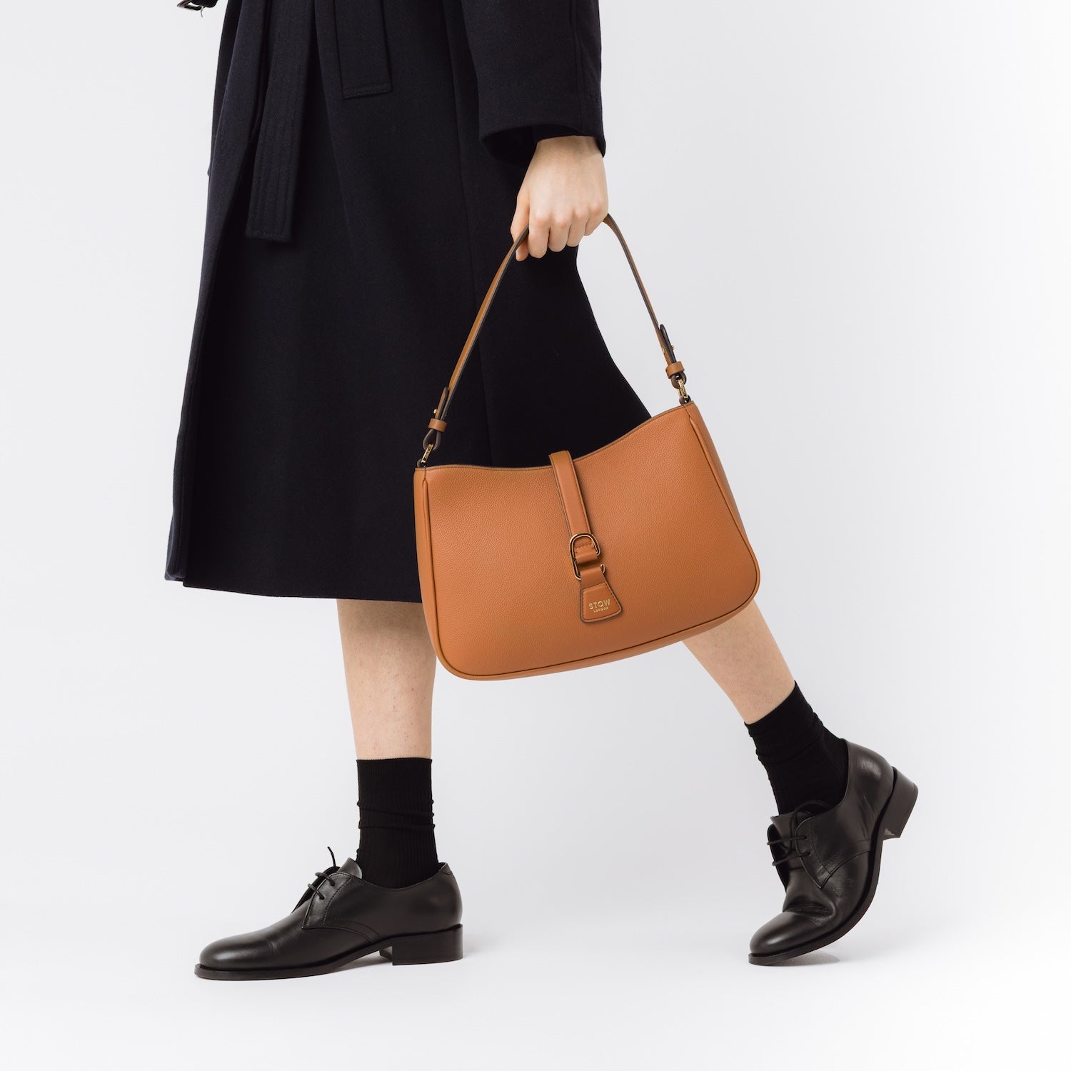 Model holding STOW London's Beth bag in Earth Tan pebbled leather at her side featuring gold-coated stainless steel hardware on a white background