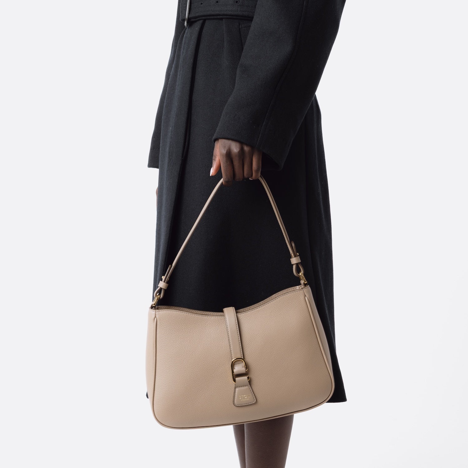Model holding STOW London's Beth bag in Warm Taupe pebbled and smooth leather at her side featuring gold-coated stainless steel hardware and clip on a white background