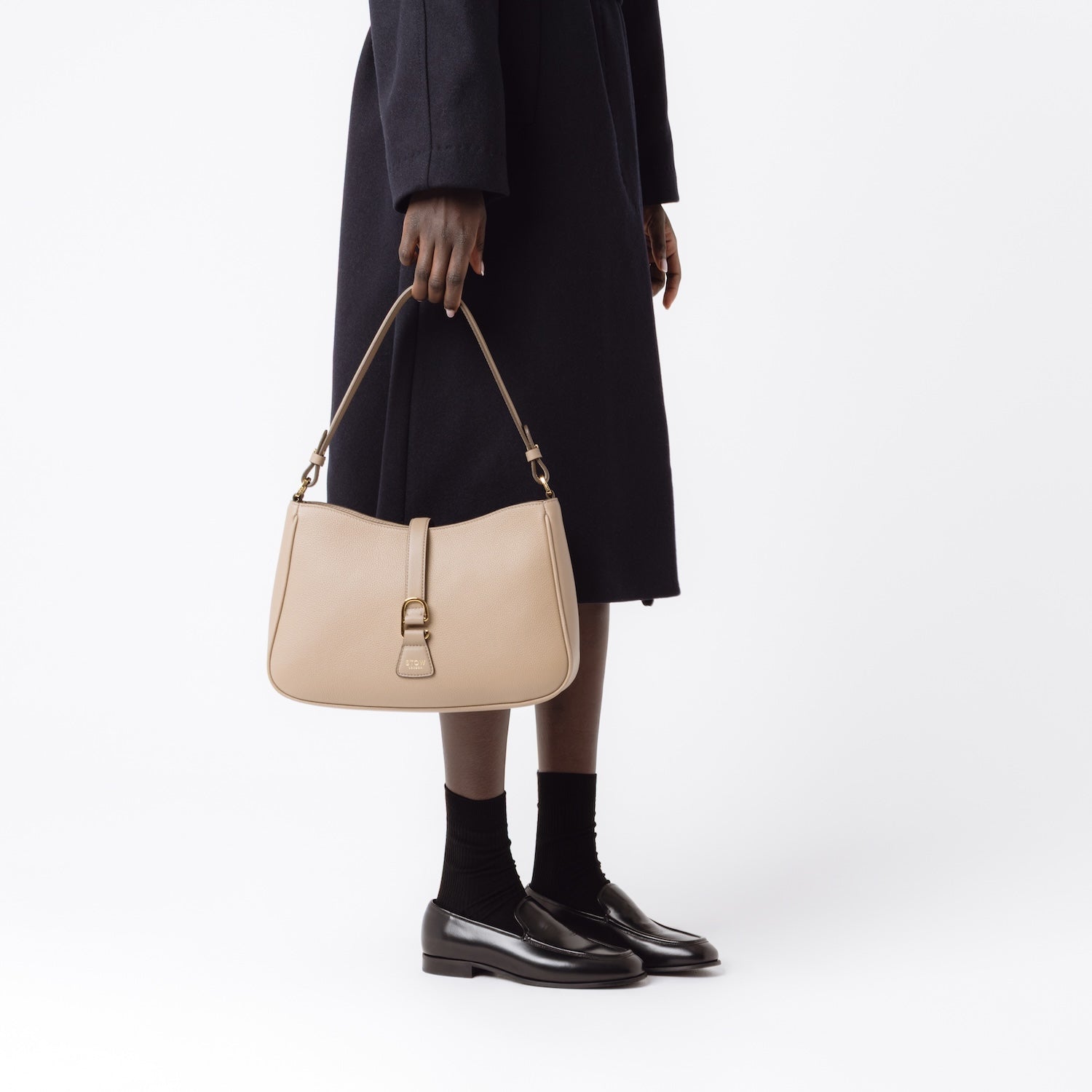 Model holding STOW London's Beth bag in Warm Taupe pebbled and smooth leather at her side featuring gold-coated stainless steel hardware and clip on a white background
