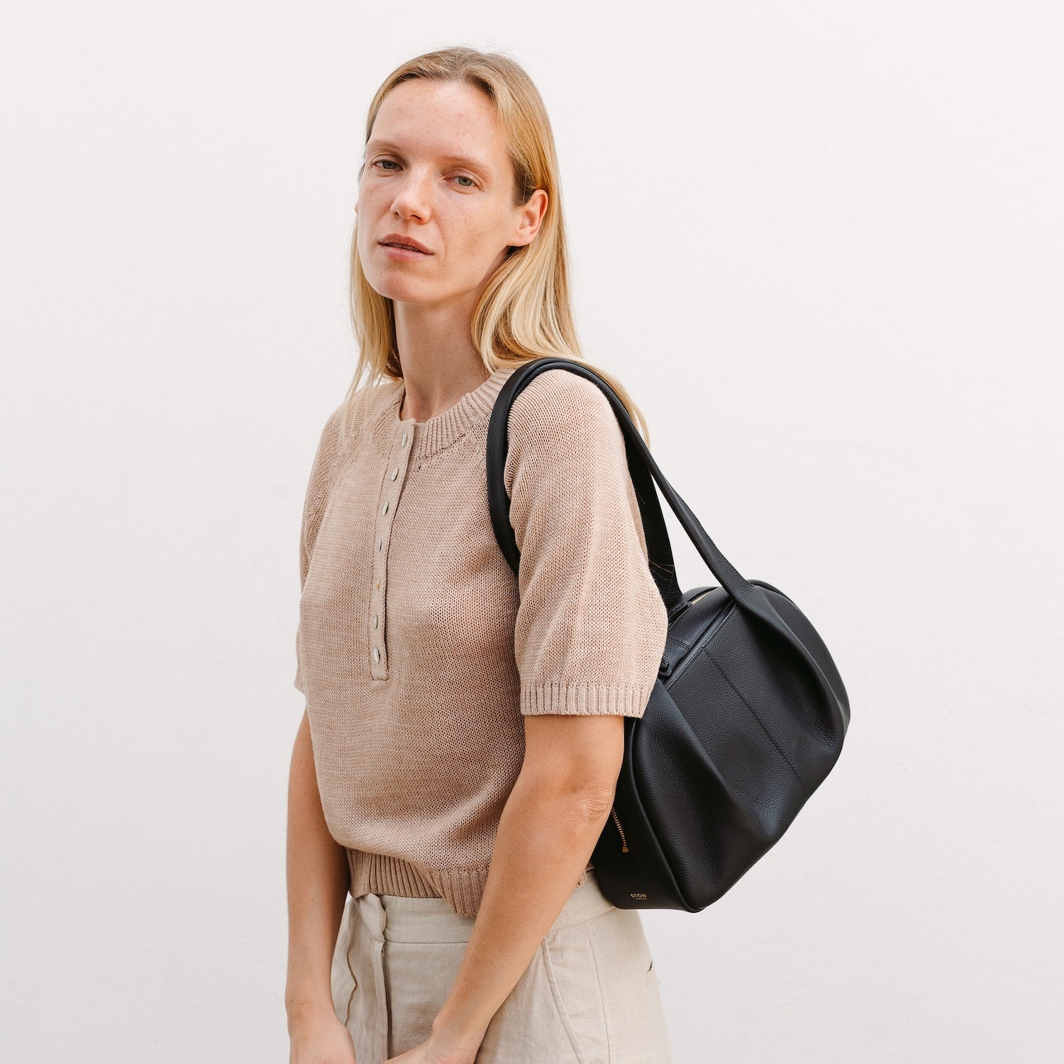 model wearing STOW London's Bowling bag in Black pebbled leather on her shoulder featuring the pleated front panel and gold-coated stainless steel hardware