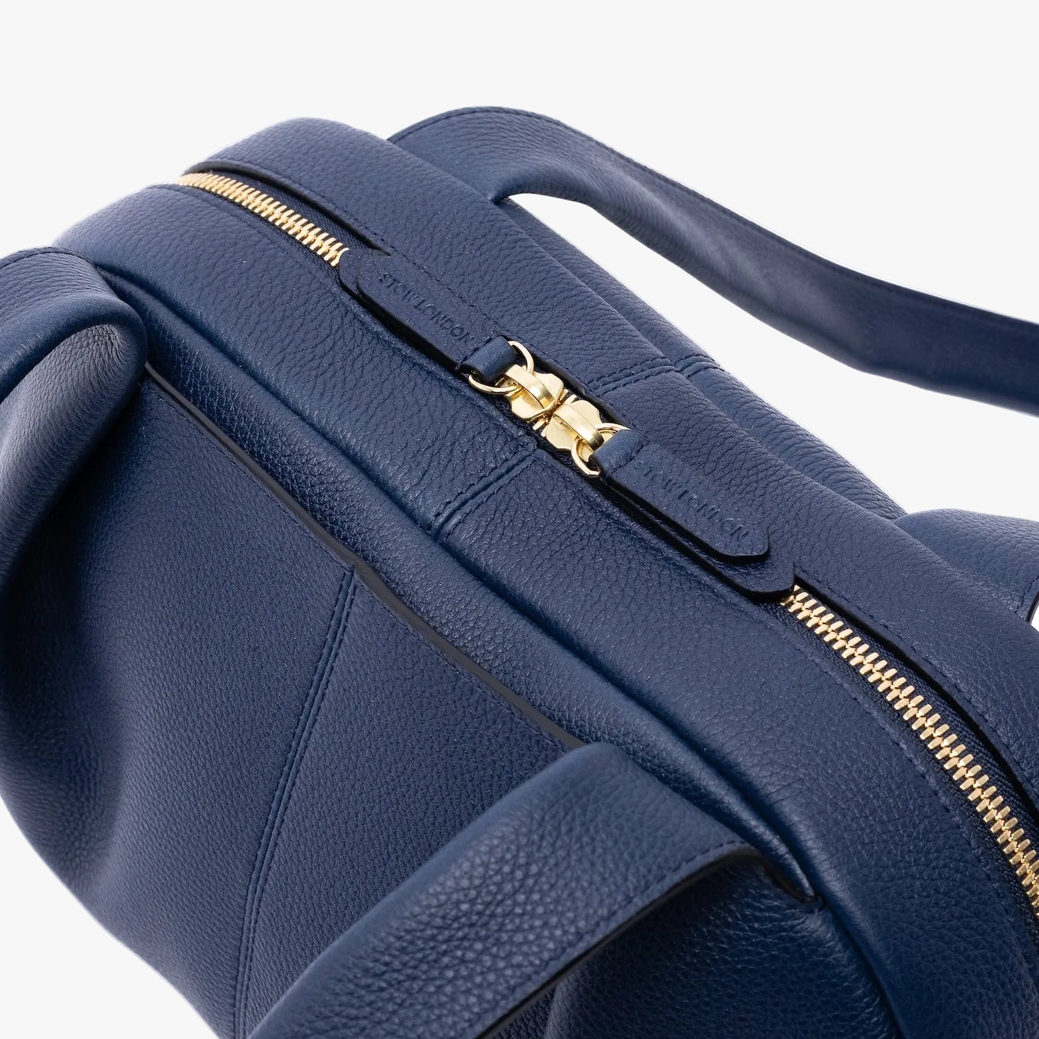 Close up of the zip top closure of STOW London's Bowling bag in Navy pebbled leather featuring gold-coated stainless steel hardware on a light grey background