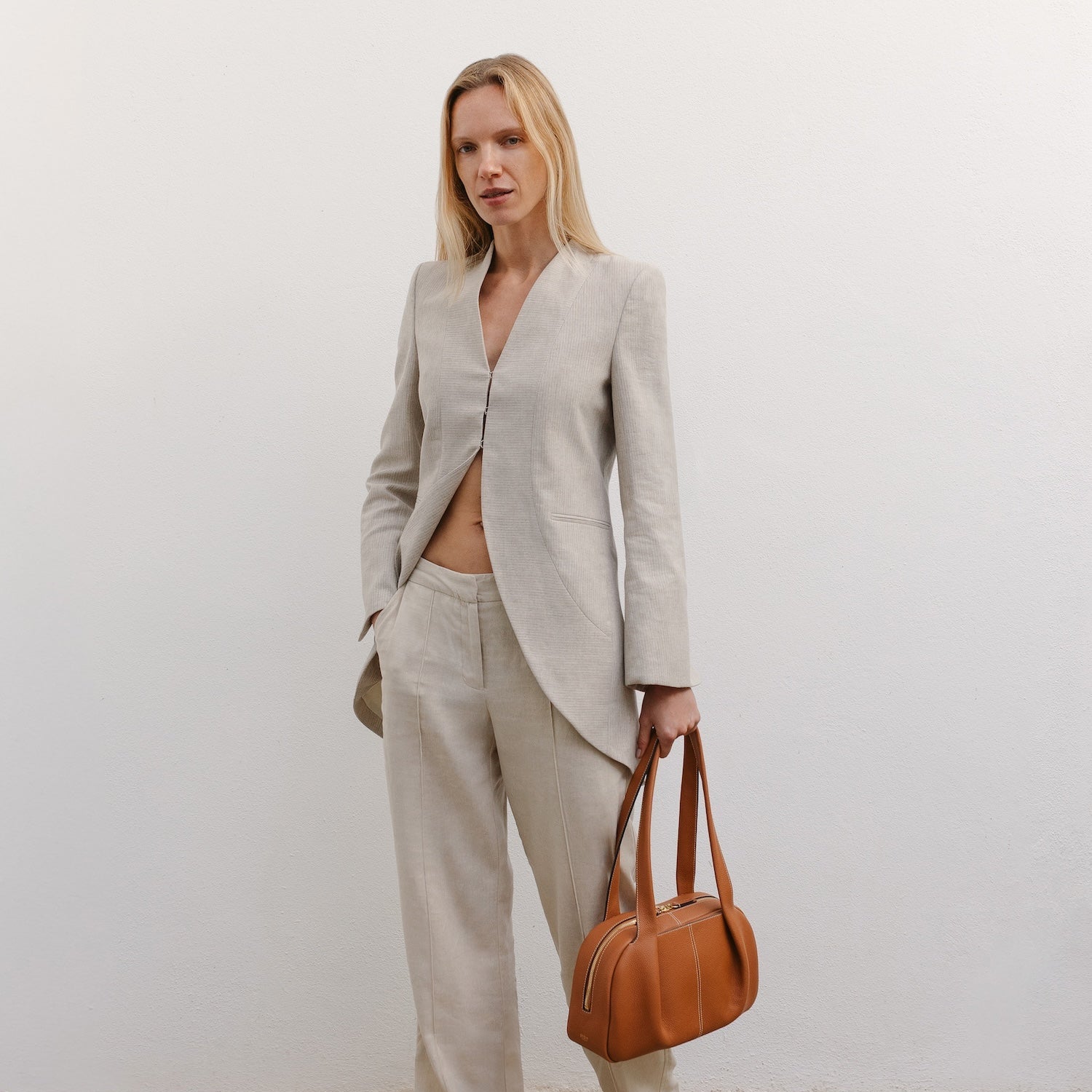 Model holding STOW London's Bowling bag in Earth Tan pebbled leather featuring the pleated front panel and gold-coated stainless steel hardware