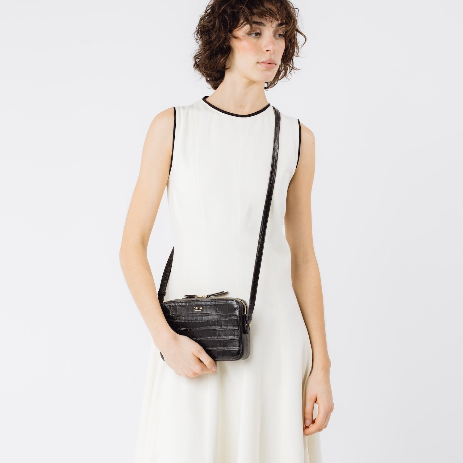 Model wearing STOW London's Camera bag in Black Embossed Croc leather crossbody with adjustable strap featuring gold-coated stainless steel hardware on a white background