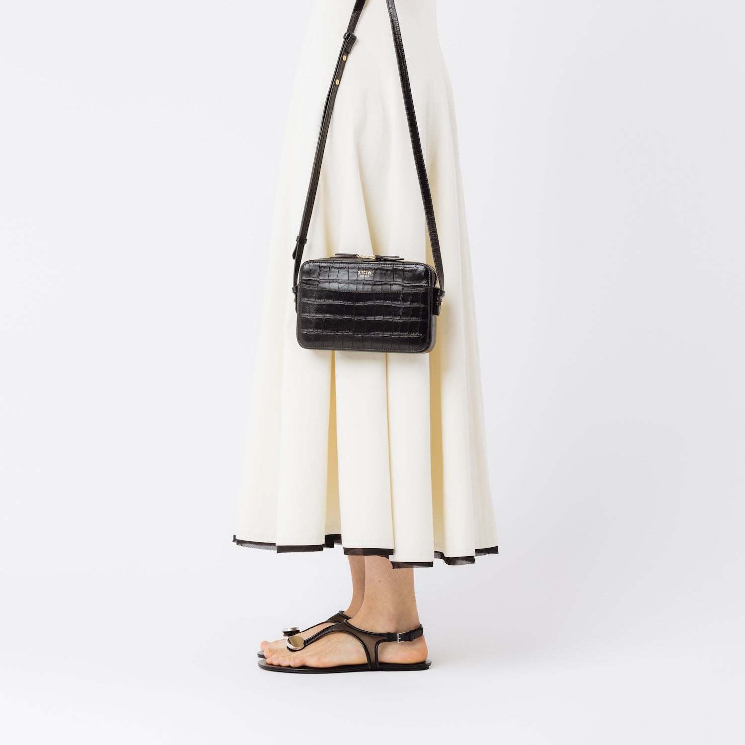 Model holding STOW London's Camera bag in Black Embossed Croc leather with detachable crossbody strap featuring gold-coated stainless steel hardware on a white background