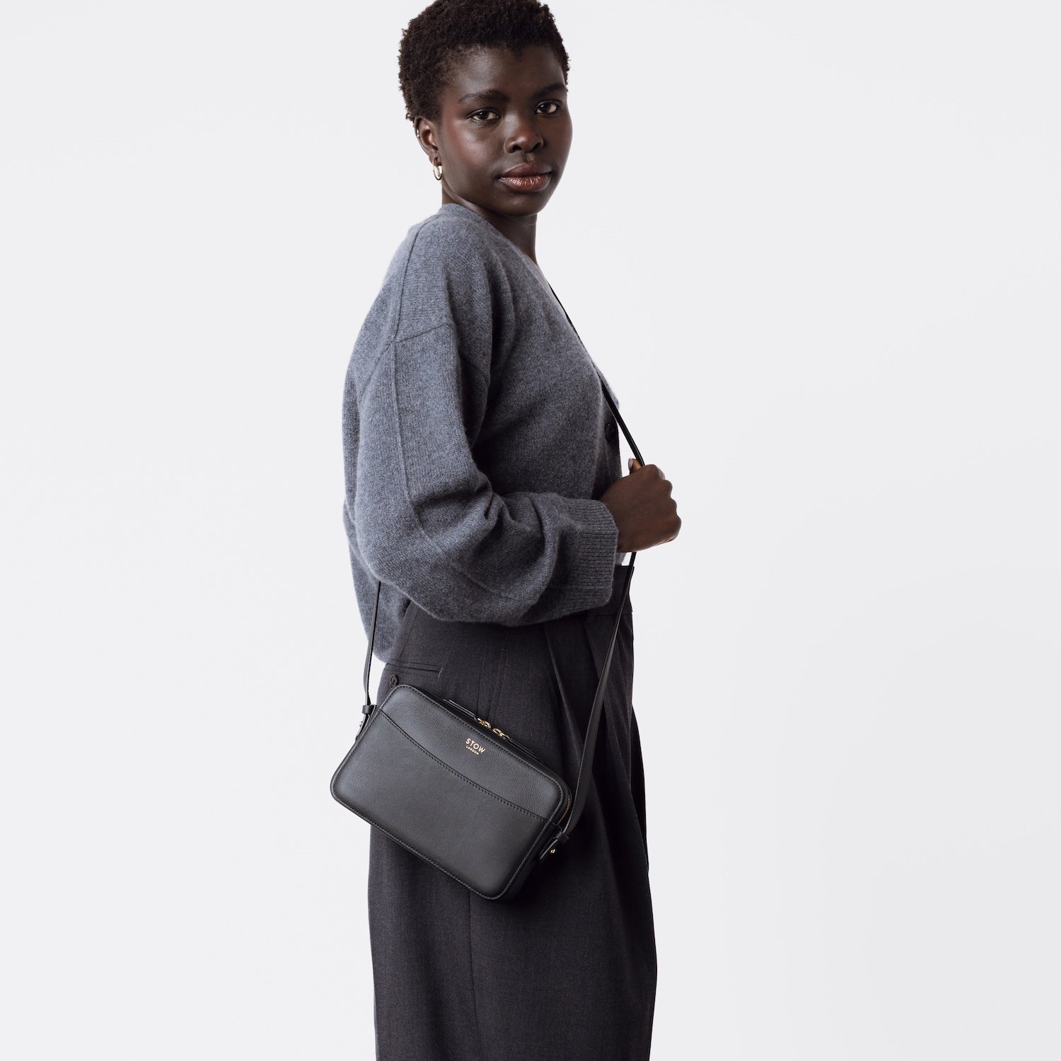 Model wearing STOW London's Camera bag in Black smooth leather crossbody with adjustable strap featuring gold-coated stainless steel hardware on a white background