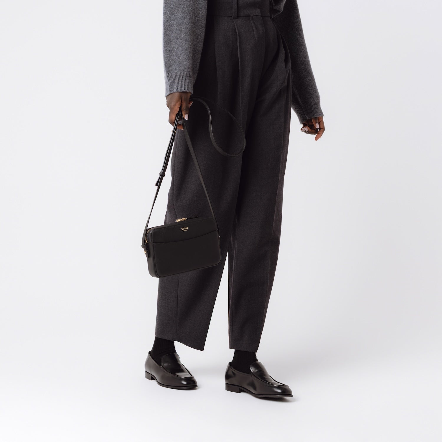 Model holding STOW London's Camera bag in Black smooth leather by its detachable leather strap at her side featuring gold-coated stainless steel hardware on a white background