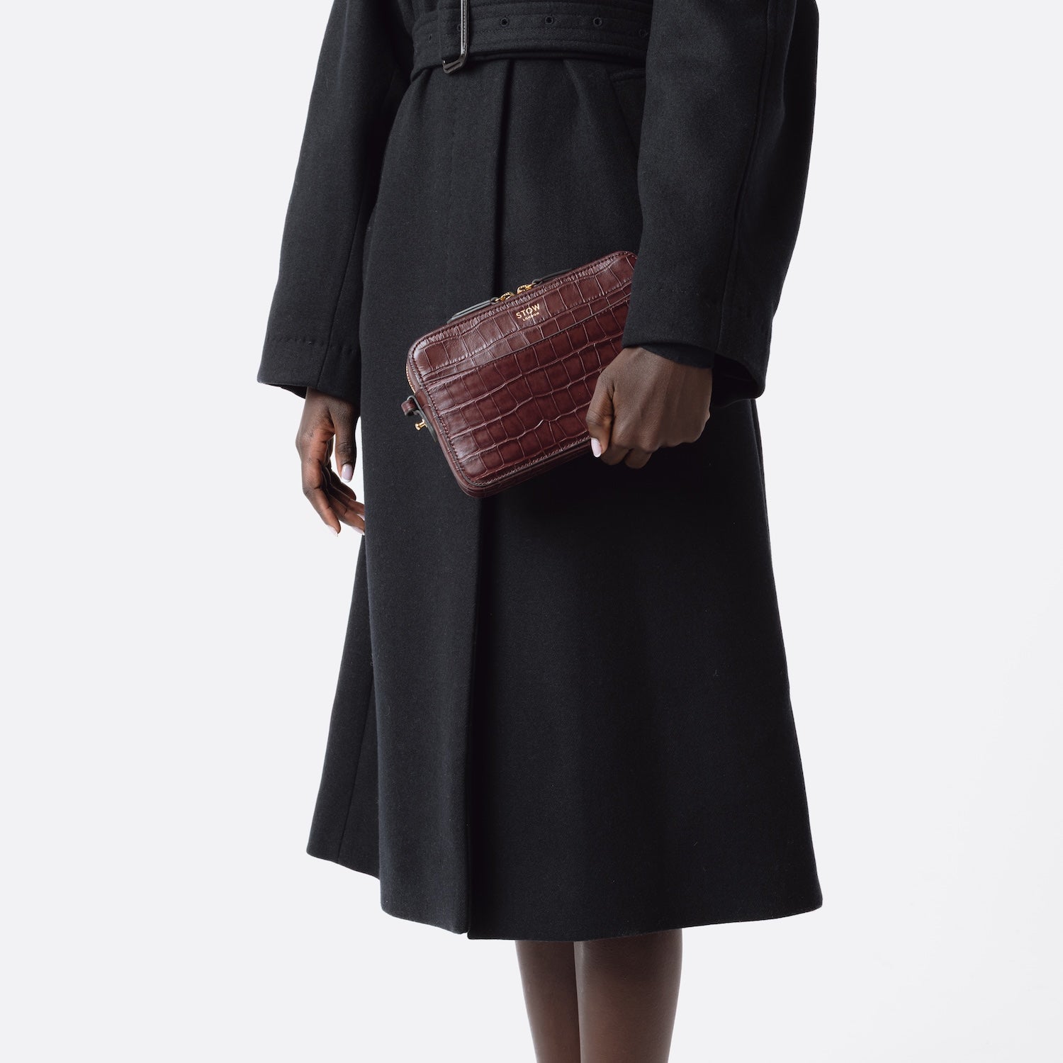 Model holding STOW London's Camera bag in Burgundy Embossed Croc leather in hand by her side featuring gold-coated stainless steel hardware on a white background