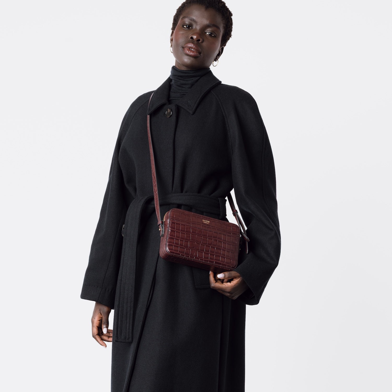 Model wearing STOW London's Camera bag in Burgundy Embossed Croc leather crossbody with adjustable strap featuring gold-coated stainless steel hardware on a white background