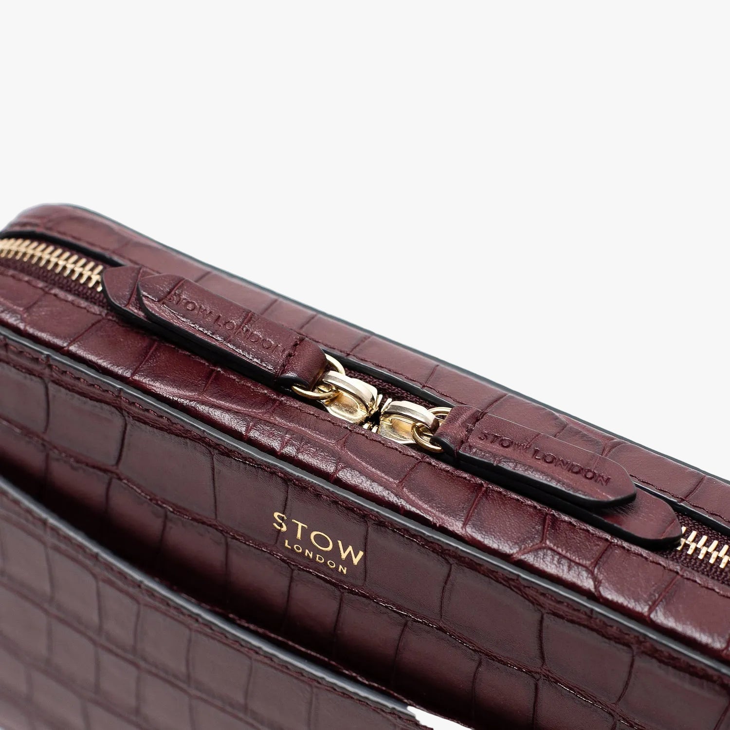 Close up of STOW London's Camera bag in Burgundy Embossed Croc leather featuring gold-coated stainless steel hardware and brand engraved pull tabs on a white background