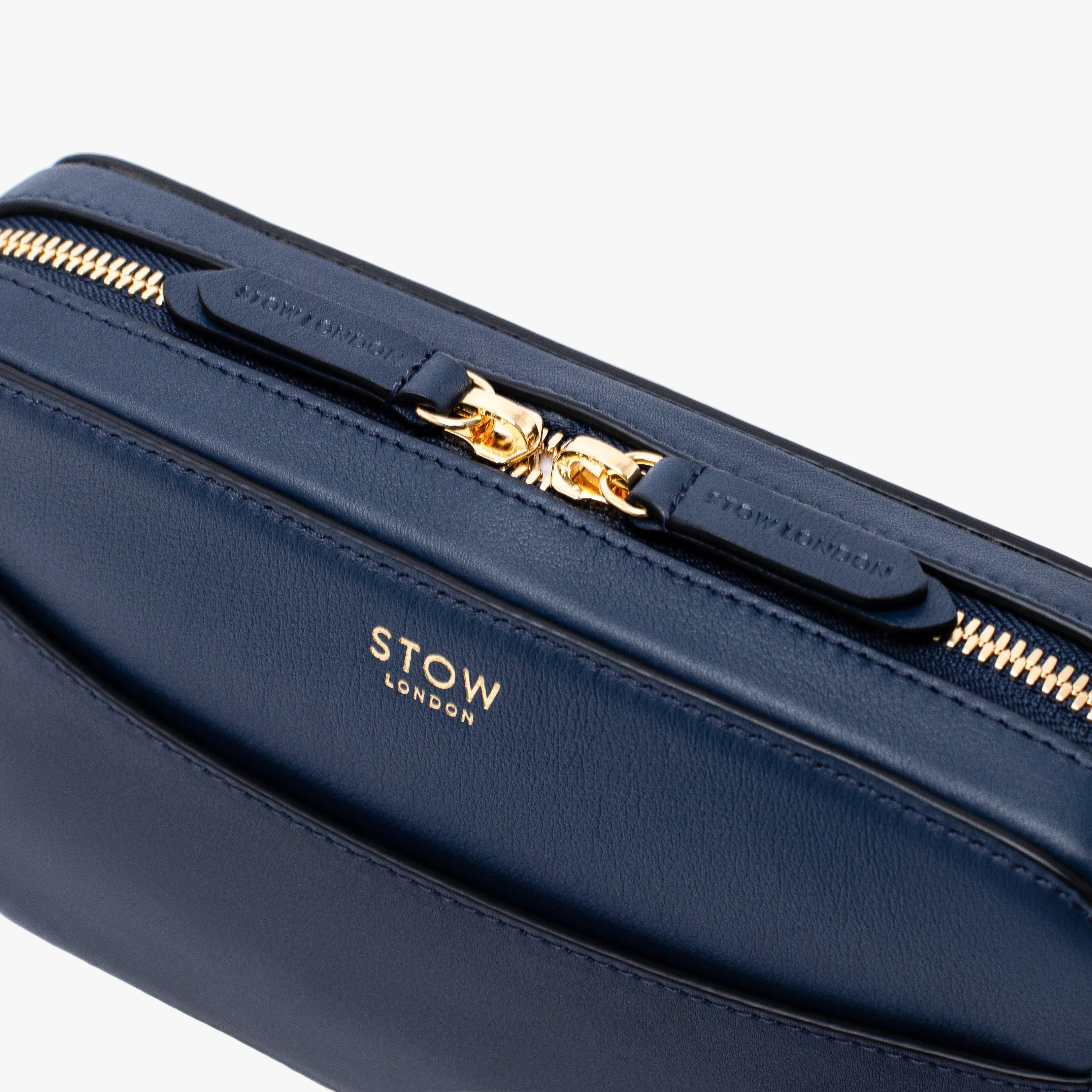 Close up of STOW London's Camera bag in Navy Smooth leather featuring gold-coated stainless steel hardware and brand engraved pull tabs on a white background