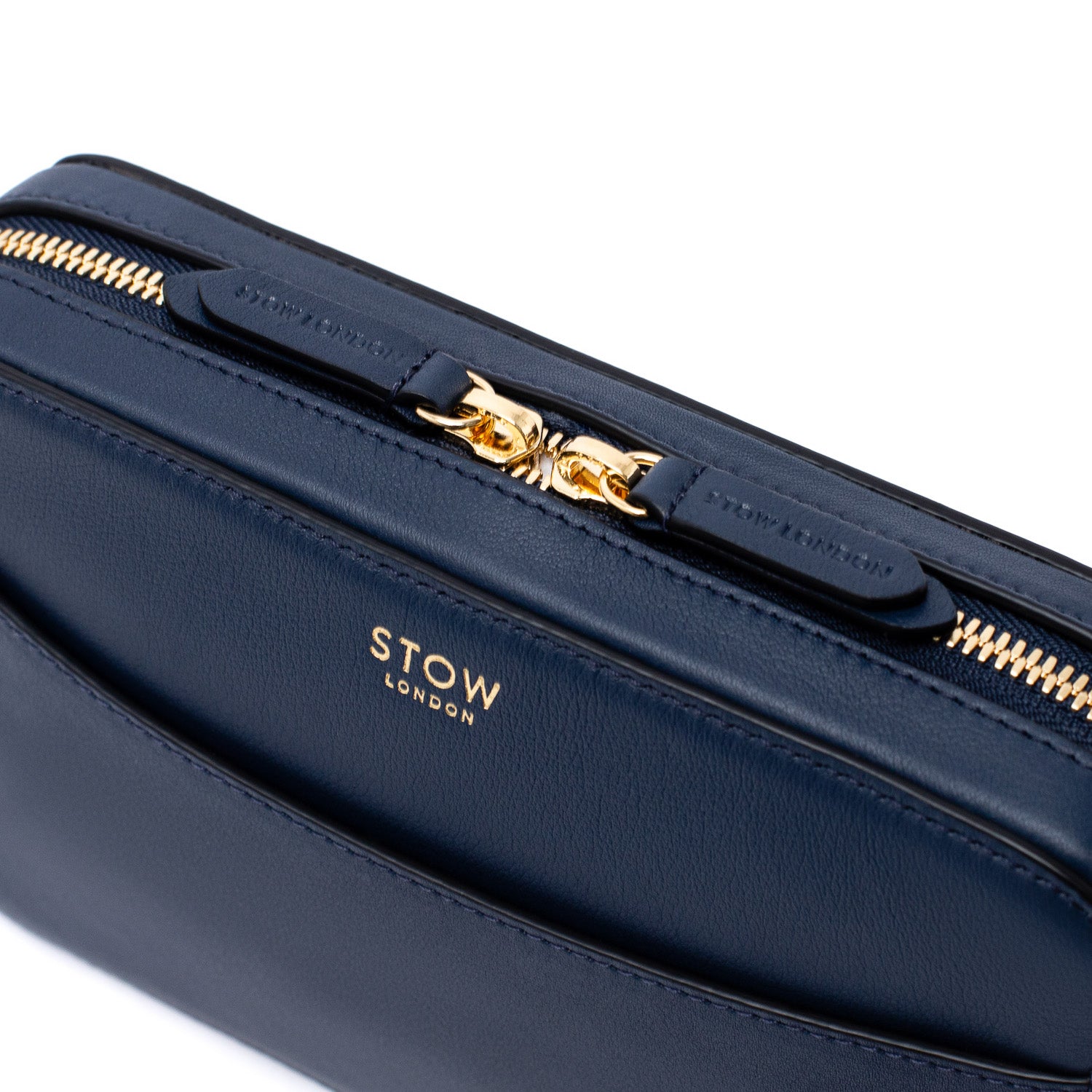 Close up of STOW London's Camera bag in Navy Smooth leather featuring gold-coated stainless steel hardware and brand engraved pull tabs on a white background