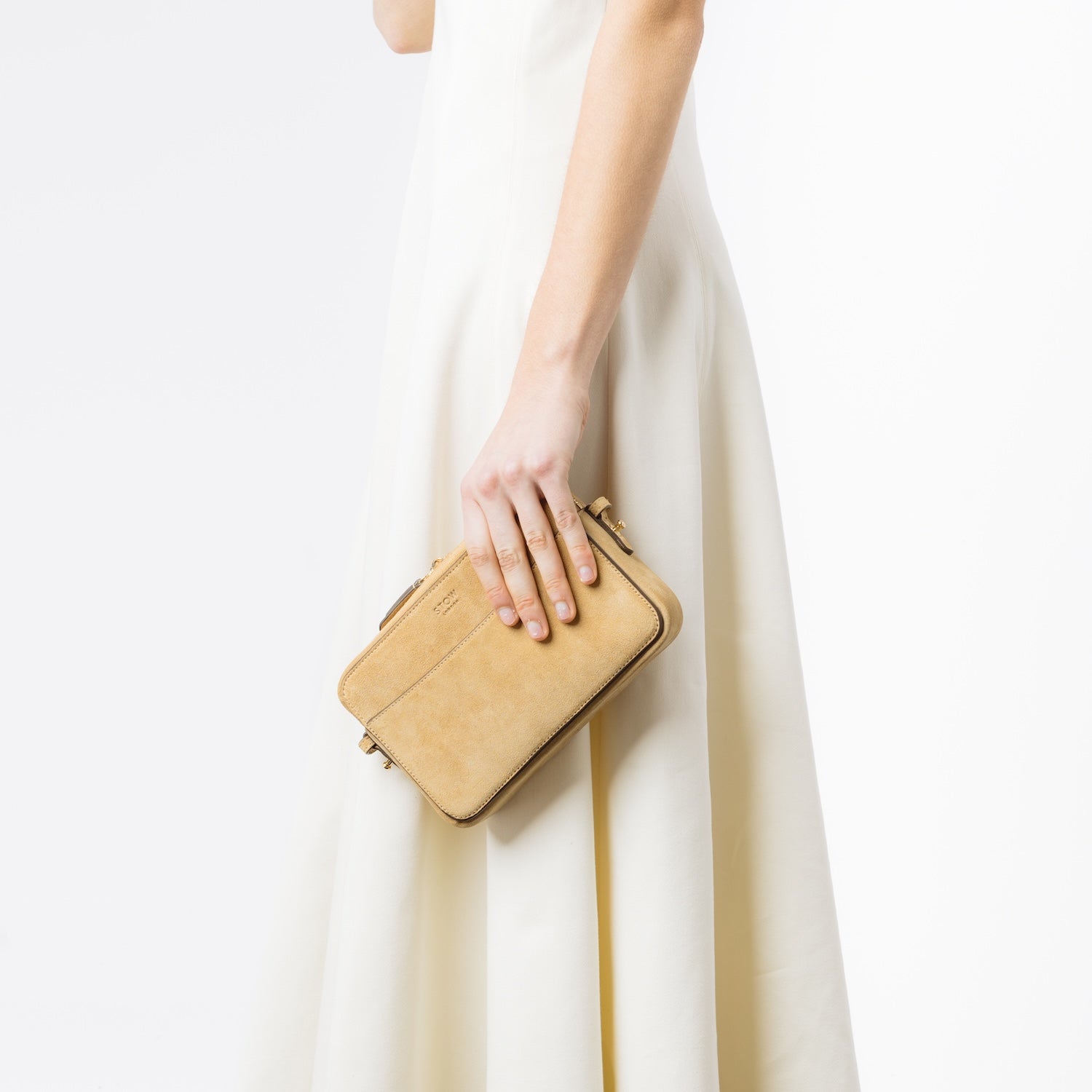 Model holding STOW London's Camera bag in Soft Sand Suede leather in hand at her side featuring gold-coated stainless steel hardware on a white background