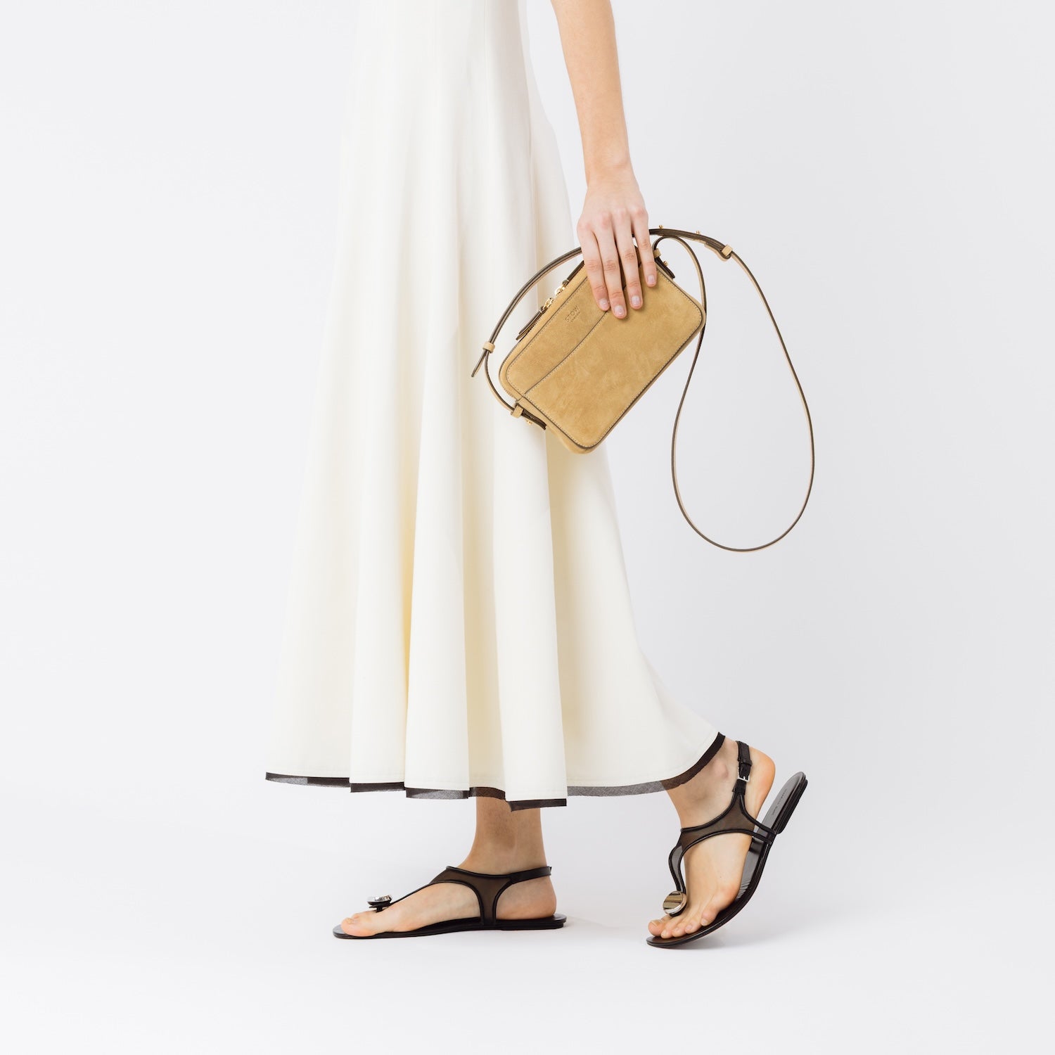 Model holding STOW London's Camera bag in Soft Sand Suede leather in her hand at her side featuring gold-coated stainless steel hardware on a white background