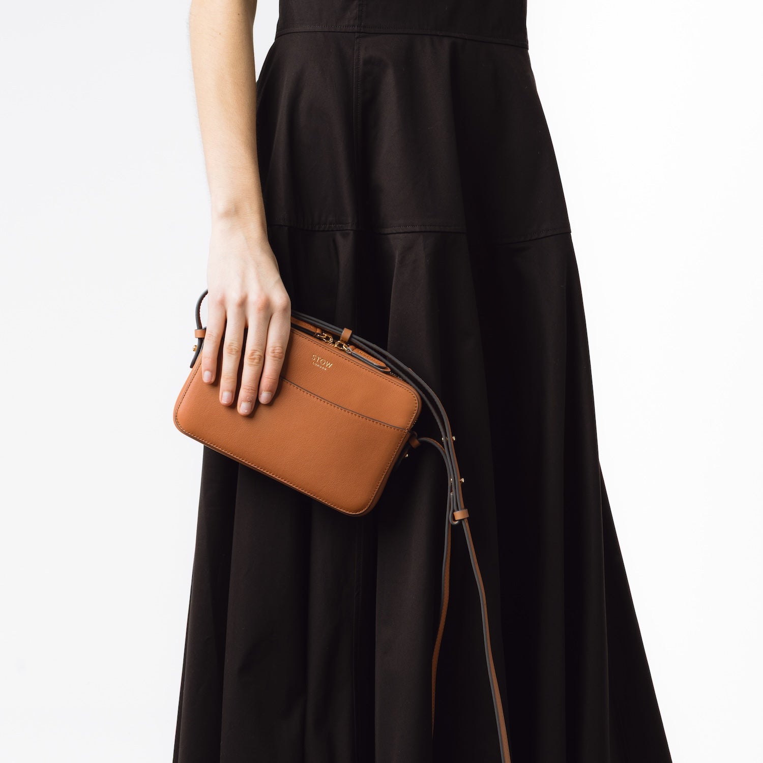 STOW London's Camera bag in Earth Tan smooth leather with detachable crossbody strap held by model at her side featuring gold-coated stainless steel hardware on a white background