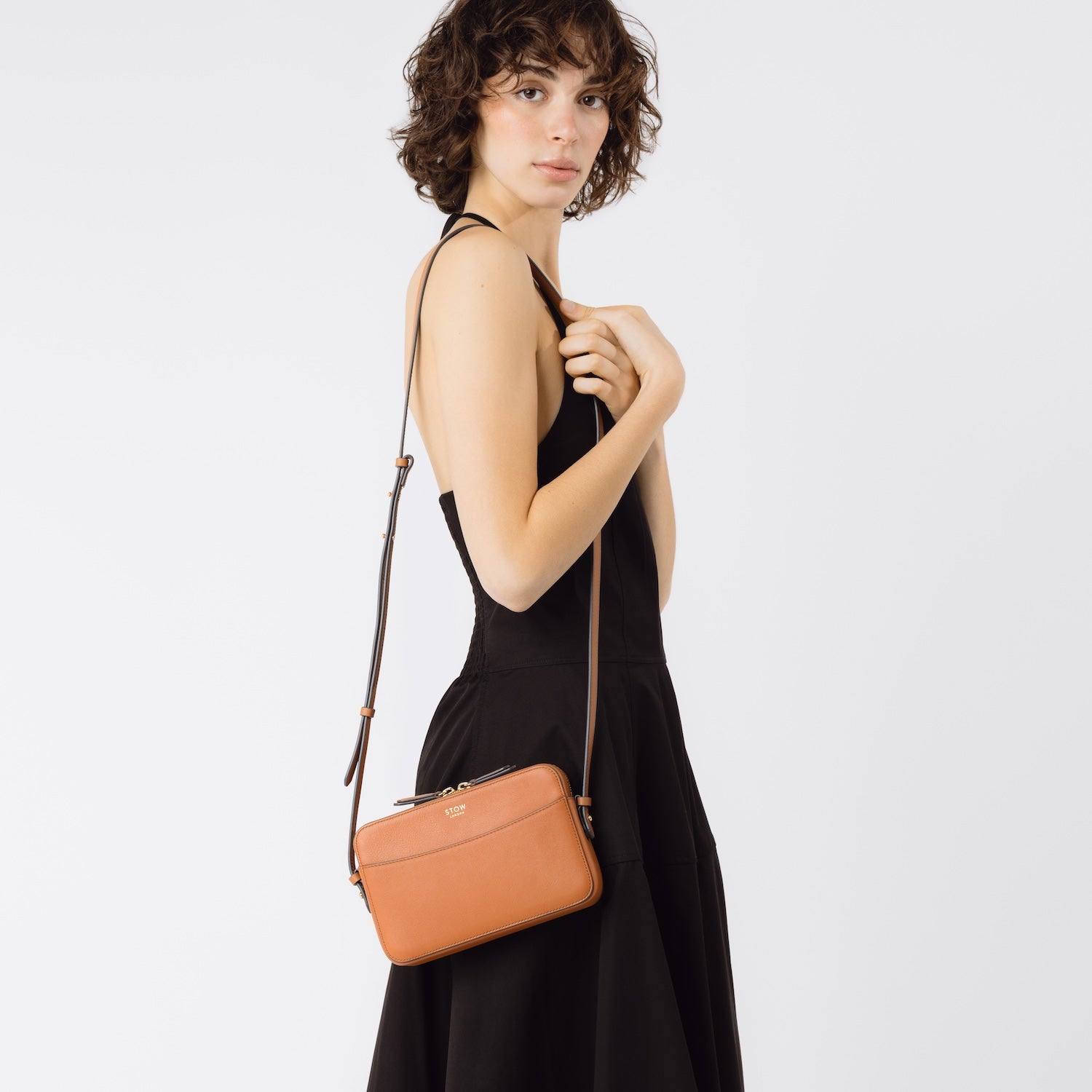 STOW London's Camera bag in Earth Tan smooth leather worn over model's shoulder with detachable crossbody strap featuring gold-coated stainless steel hardware on a white background