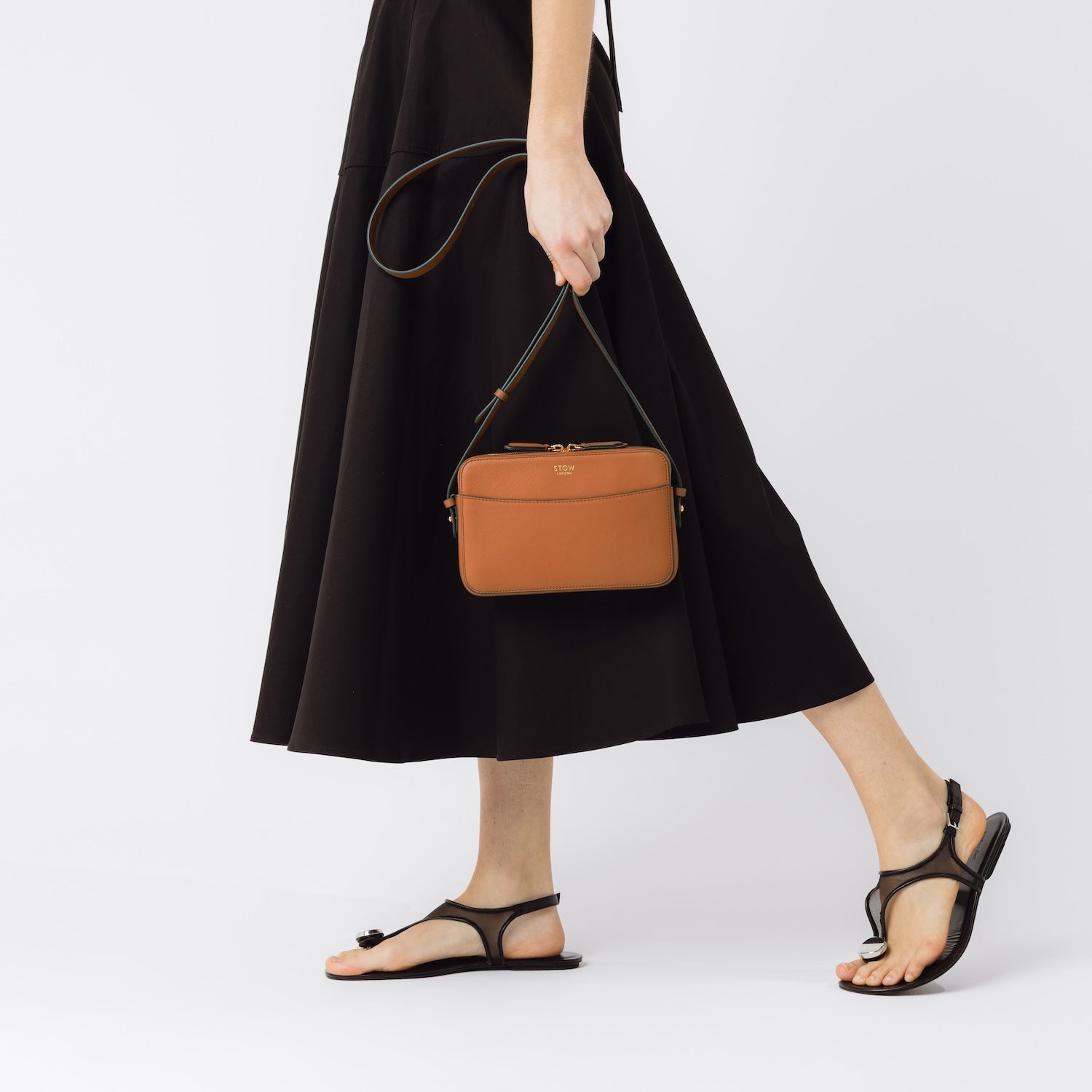 Model holding STOW London's Camera bag in Earth Tan smooth leather at her side by the crossbody strap featuring gold-coated stainless steel hardware