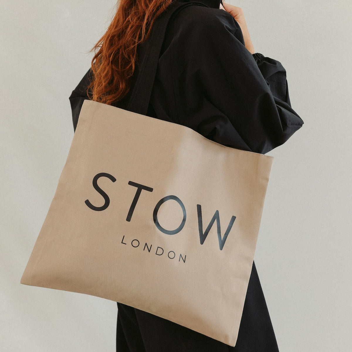 Model with long red hair carrying STOW London's Beige Canvas Tote bag featuring the brand name in black text, set against a plain light grey background.