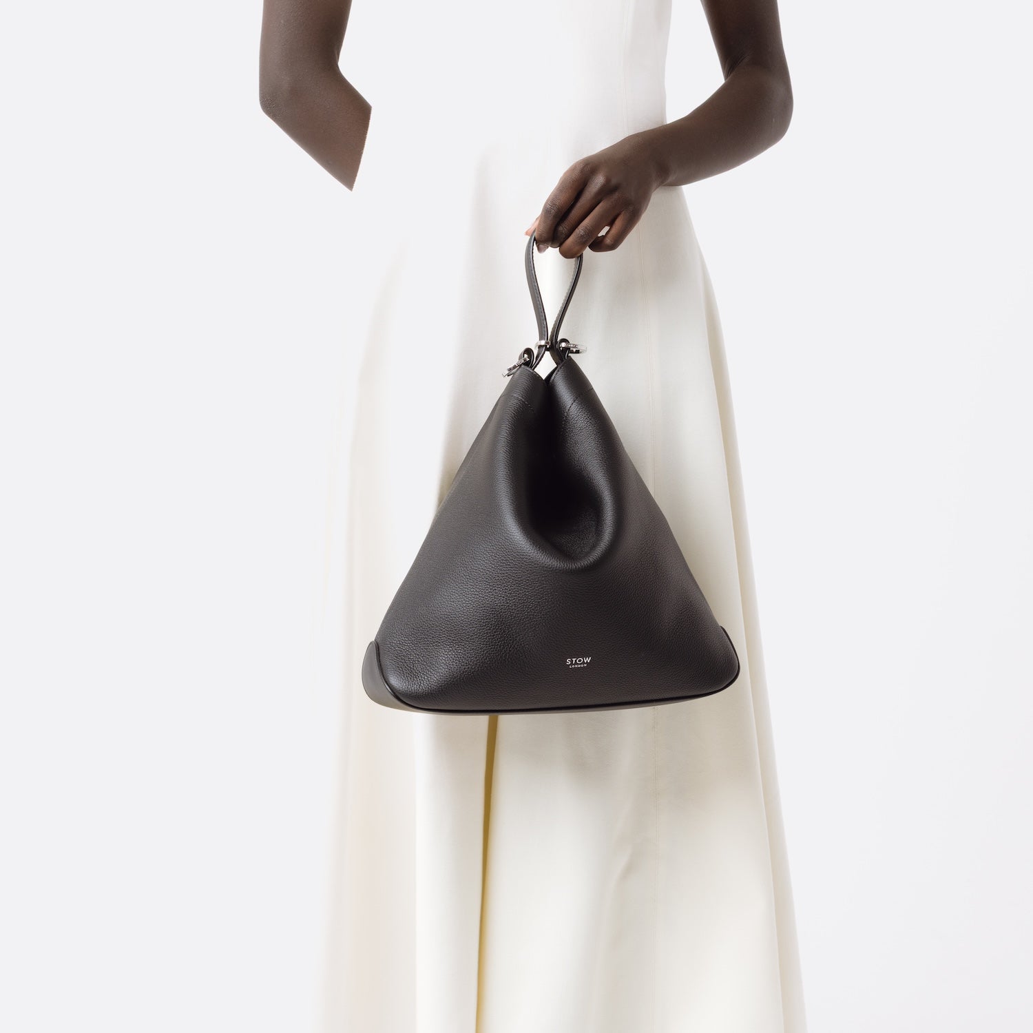 Model holding Stow London's Curve bag in Black pebbled leather at her front featuring silver stainless steel hardware on a light grey background