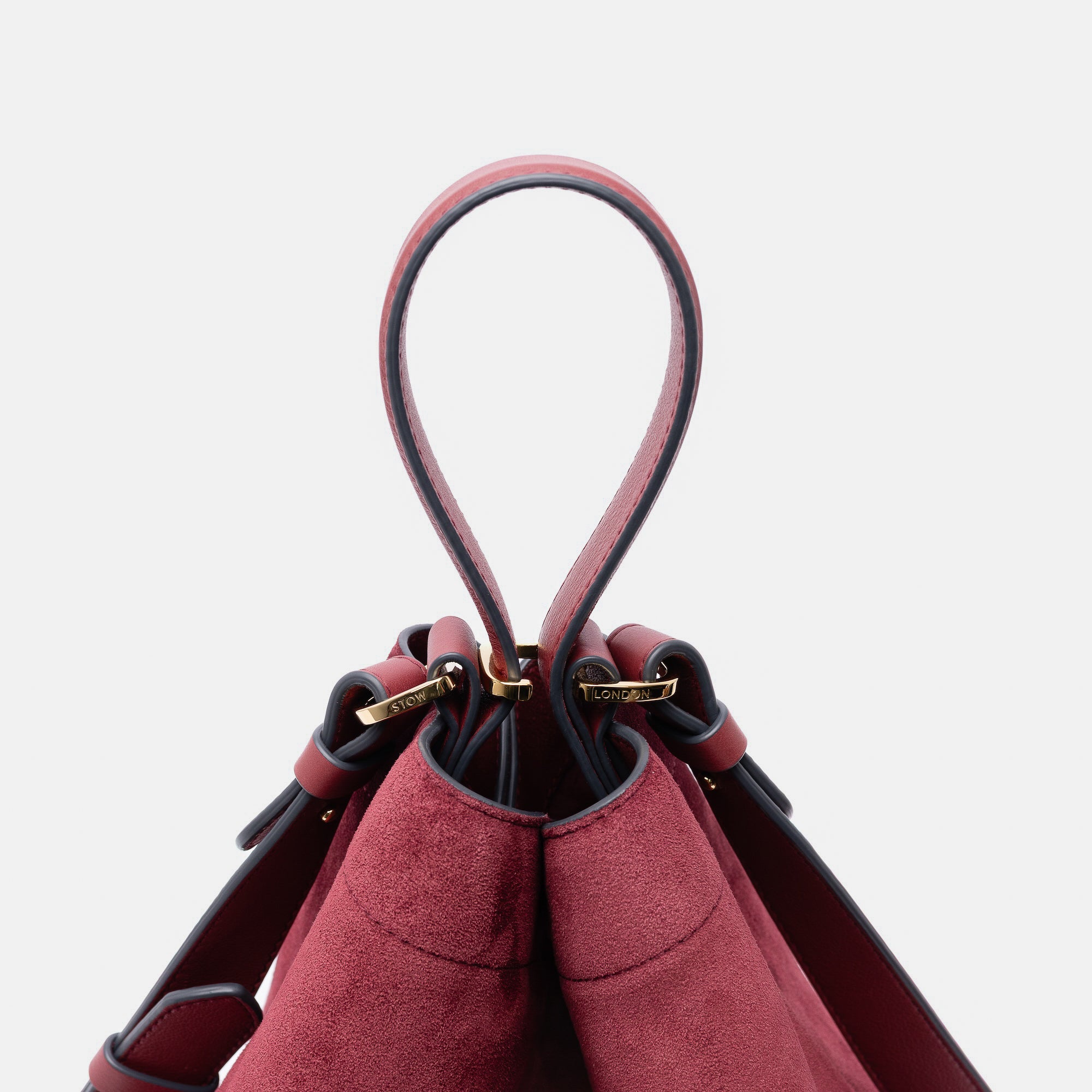 Close up of the handles on Stow London's Curve bag in Burgundy suede leather featuring gold-coated stainless steel hardware and clip and detachable strap on a light grey background