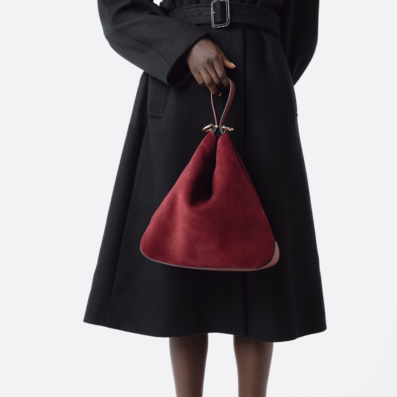 Model holding Stow London's Curve bag in Burgundy suede leather by handle at her front featuring gold-coated stainless steel hardware on a light grey background