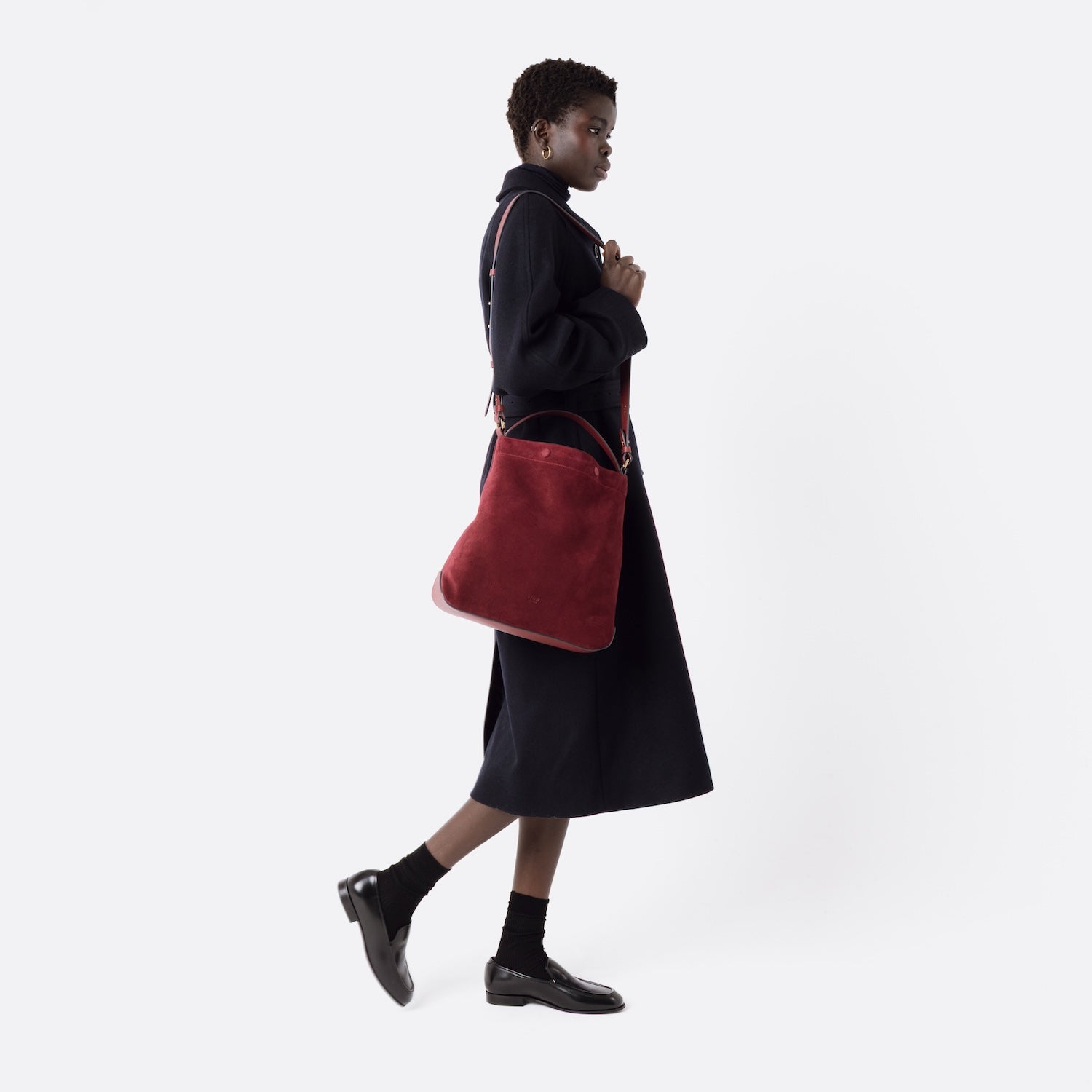 Model wearing Stow London's Curve bag in Burgundy suede leather on her shoulder featuring gold-coated stainless steel hardware on a light grey background