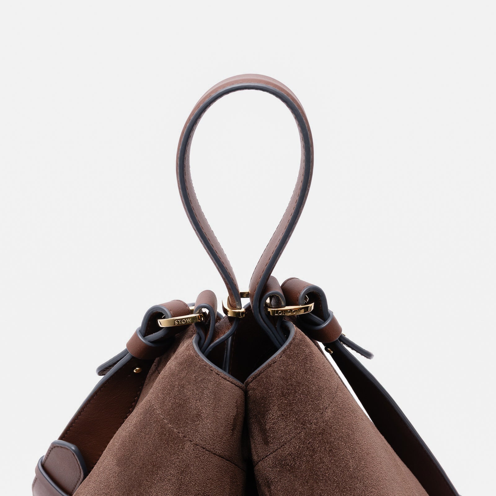 Close up of the handles on Stow London's Curve bag in Chocolate suede leather featuring gold-coated stainless steel hardware and clip and detachable strap on a light grey background
