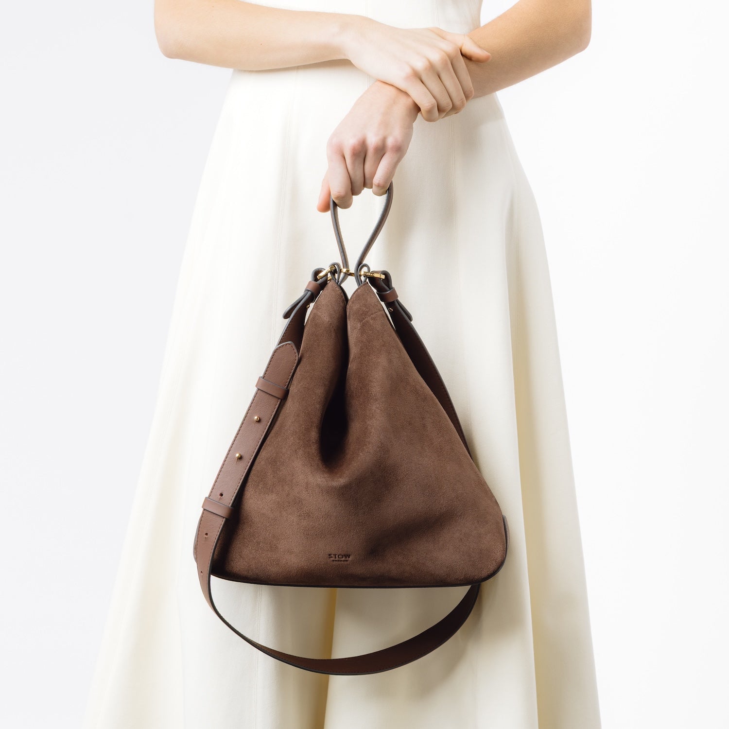 Model holding Stow London's Curve bag in Chocolate suede leather at her front featuring gold-coated stainless steel hardware on a light grey background