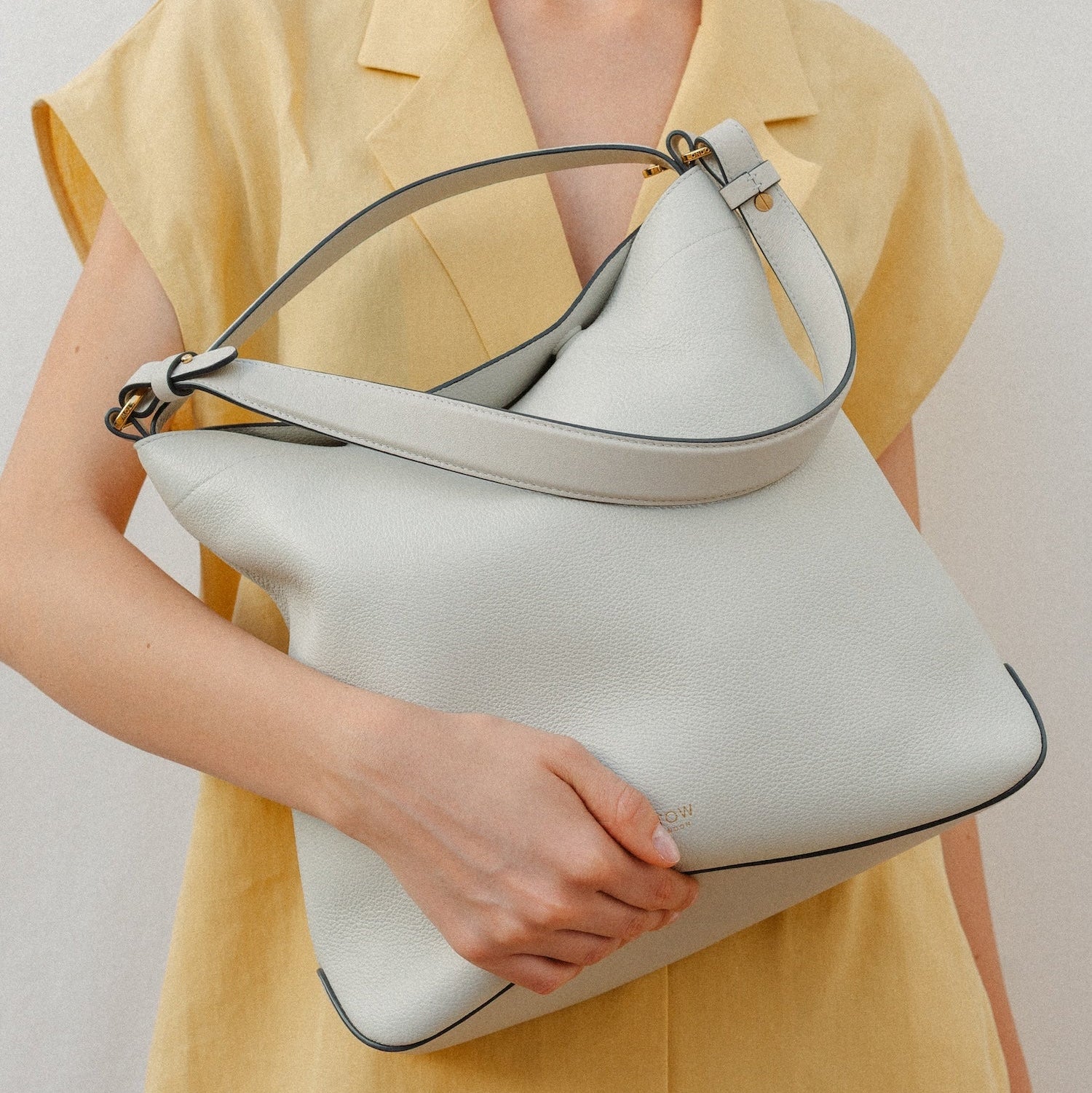 Model holding STOW London's Curve in Cloud Grey leather featuring the detachable shoulder strap and top handle with gold hardware and details