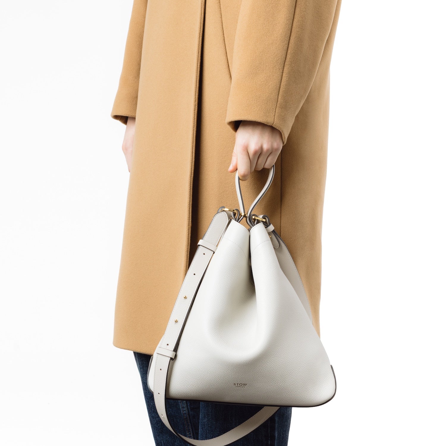 Model holding Stow London's Curve bag in Cloud Grey pebbled leather by the handle at her side in a triangular form featuring gold-coated stainless steel hardware on a light grey background