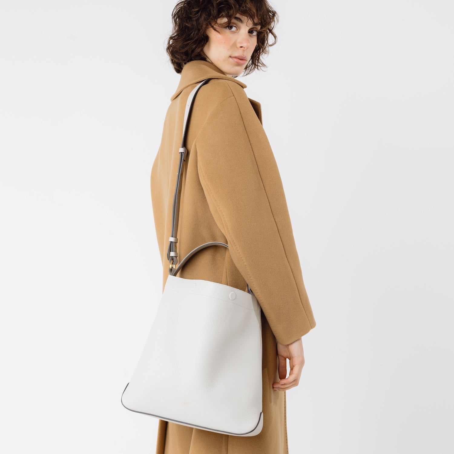 Model wearing Stow London's Curve bag in Cloud Grey pebbled leather on her shoulder featuring gold-coated stainless steel hardware on a light grey background