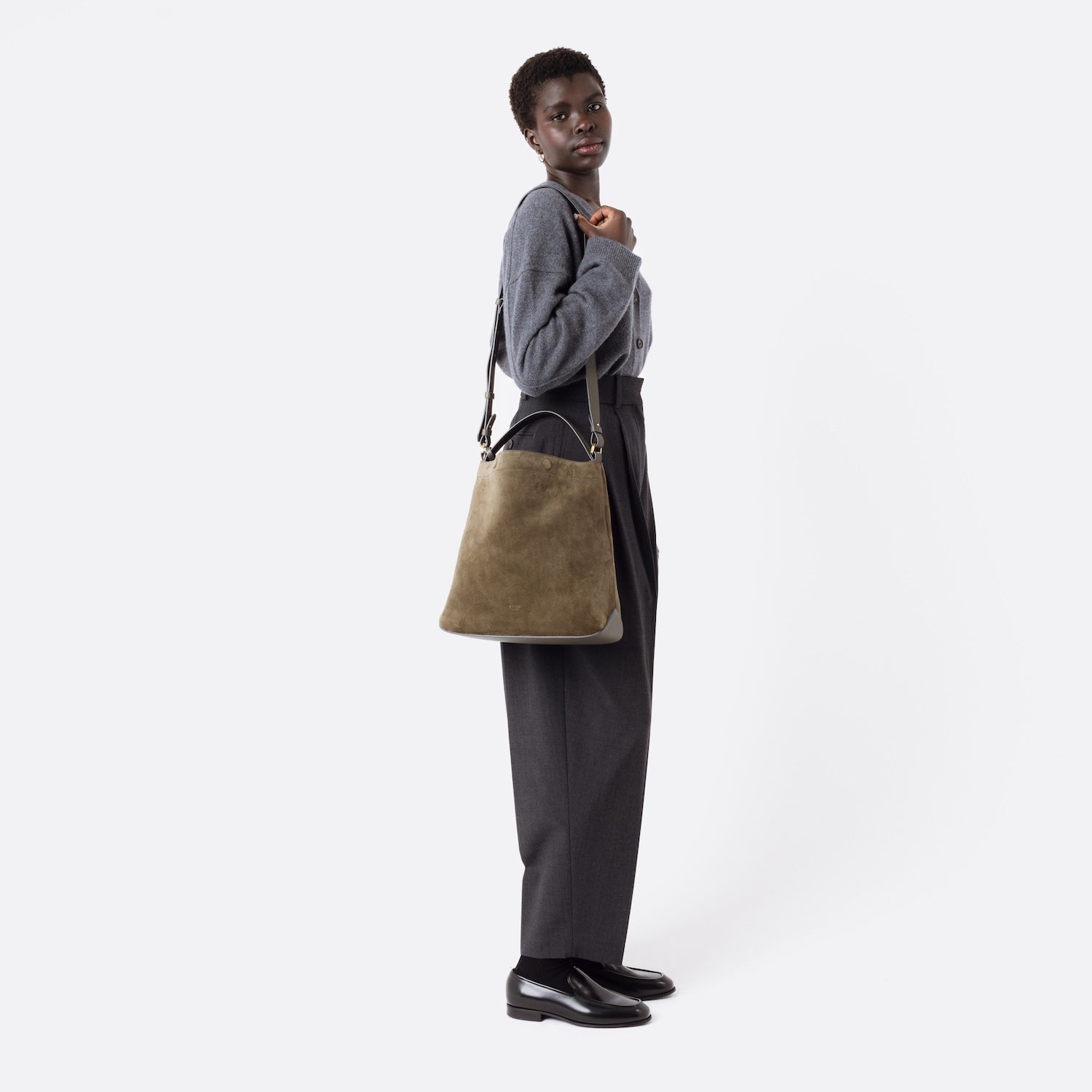 Model wearing Stow London's Curve bag in Dark Green suede leather on her shoulder featuring gold-coated stainless steel hardware and the detachable crossbody strap on a white background