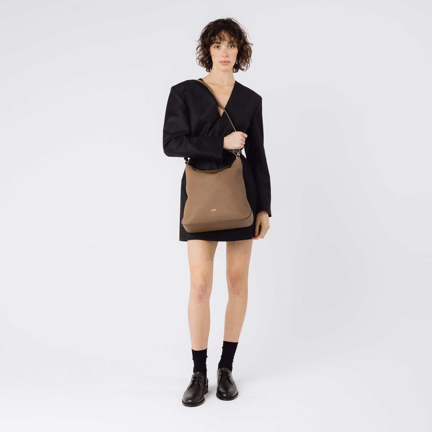 Model wearing STOW London's Curve in Mocha pebbled leather on her shoulder featuring gold-coated stainless steel hardware on a white background