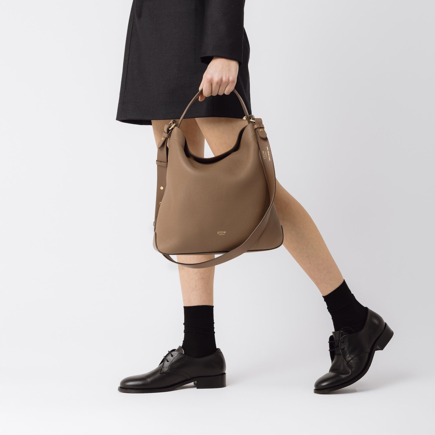 Model holding STOW London's Curve in Mocha pebbled leather by top handle at her side featuring gold-coated stainless steel hardware on a white background