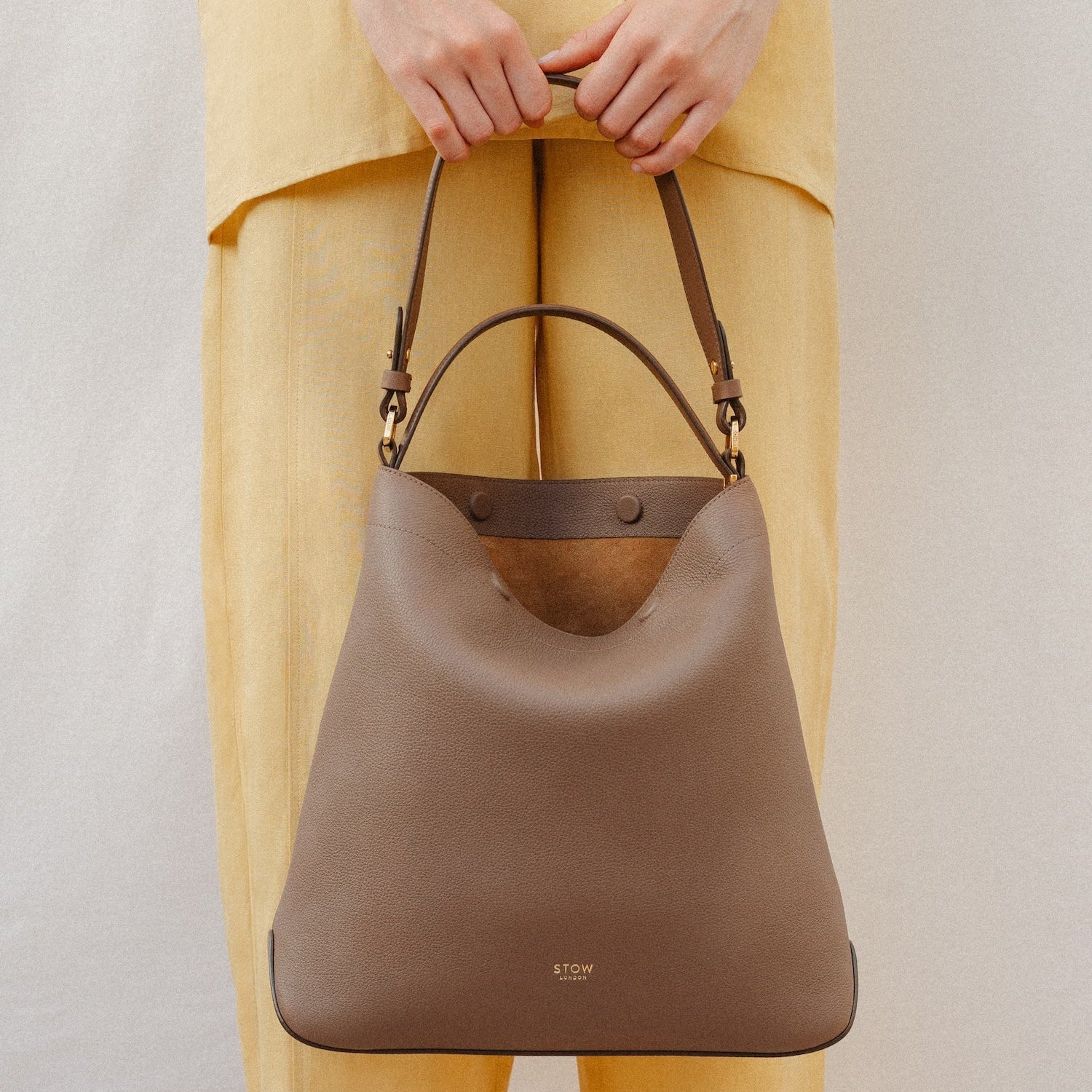 Model holding STOW London's Curve in Mocha leather featuring the detachable shoulder strap and top handle with gold hardware and details