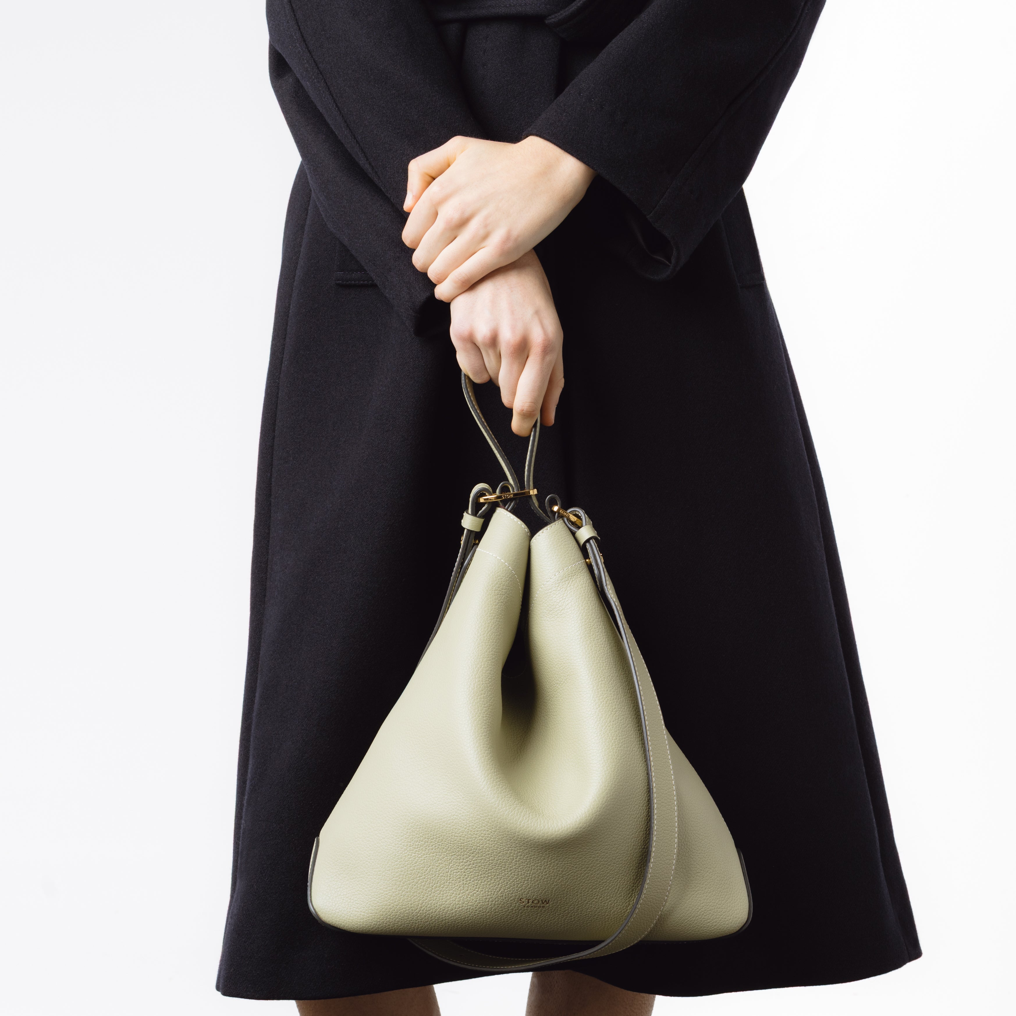 Model holding Stow London's Curve bag in Pale Green pebbled leather by the top handle featuring gold-coated stainless steel hardware on a light grey background