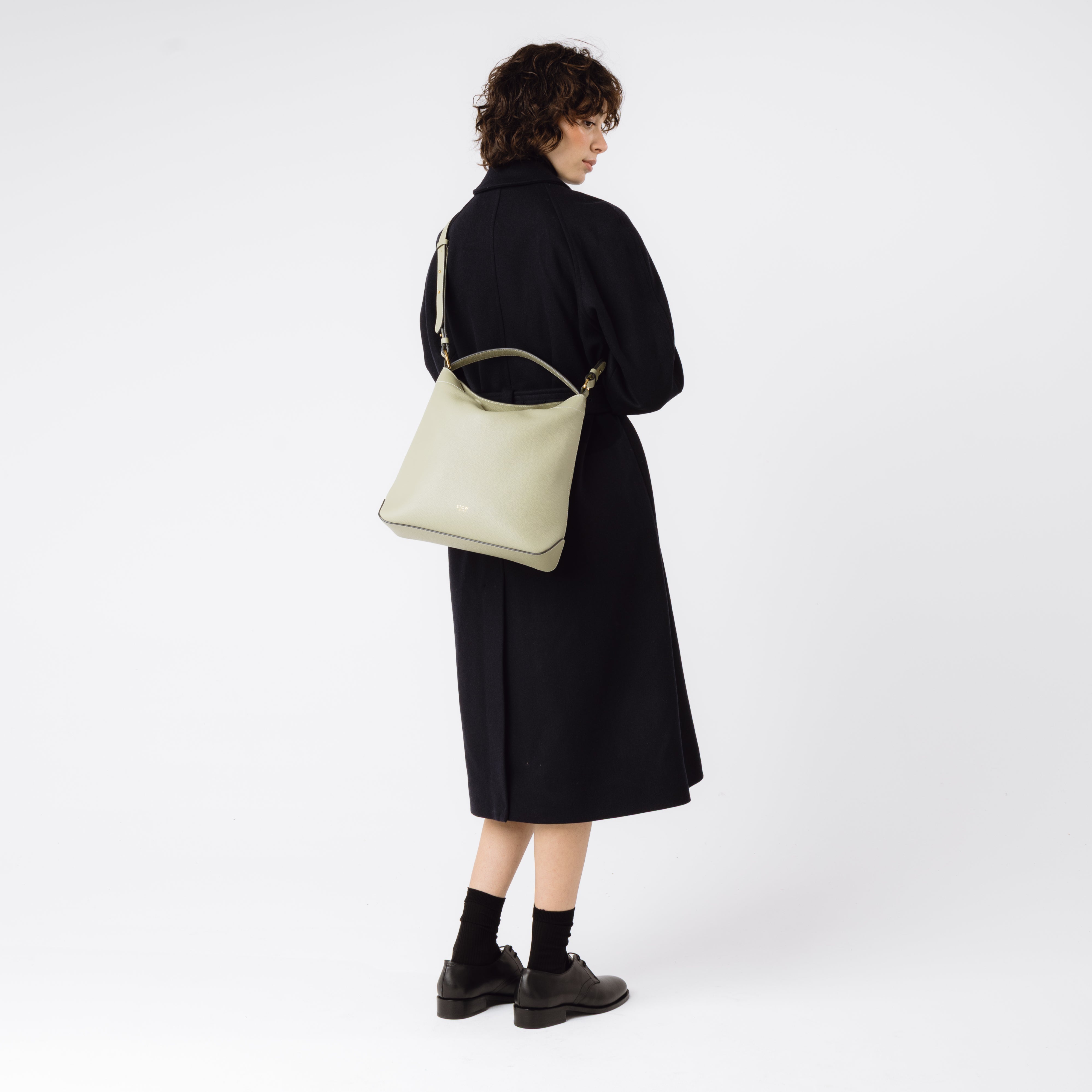 Model wearing Stow London's Curve bag in Pale Green pebbled leather crossbody featuring gold-coated stainless steel hardware on a light grey background
