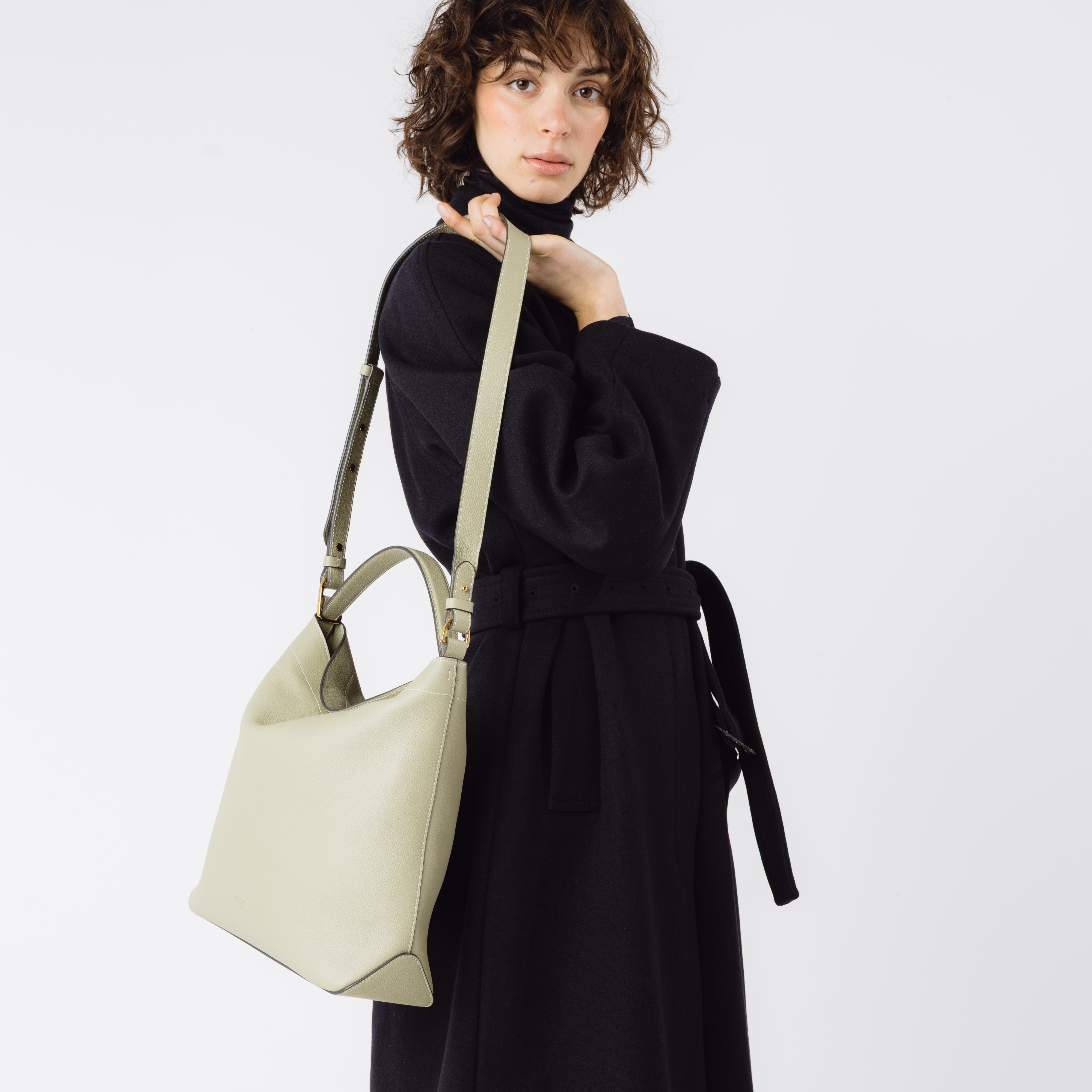 Model holding Stow London's Curve bag in Pale Green pebbled leather by the detachable crossbody strap featuring gold-coated stainless steel hardware on a light grey background