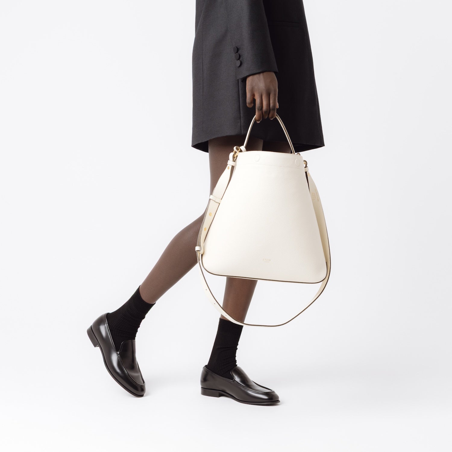 Model holding Stow London's Curve bag in Spring Moon pebbled leather at her side featuring gold-coated stainless steel hardware on a light grey background