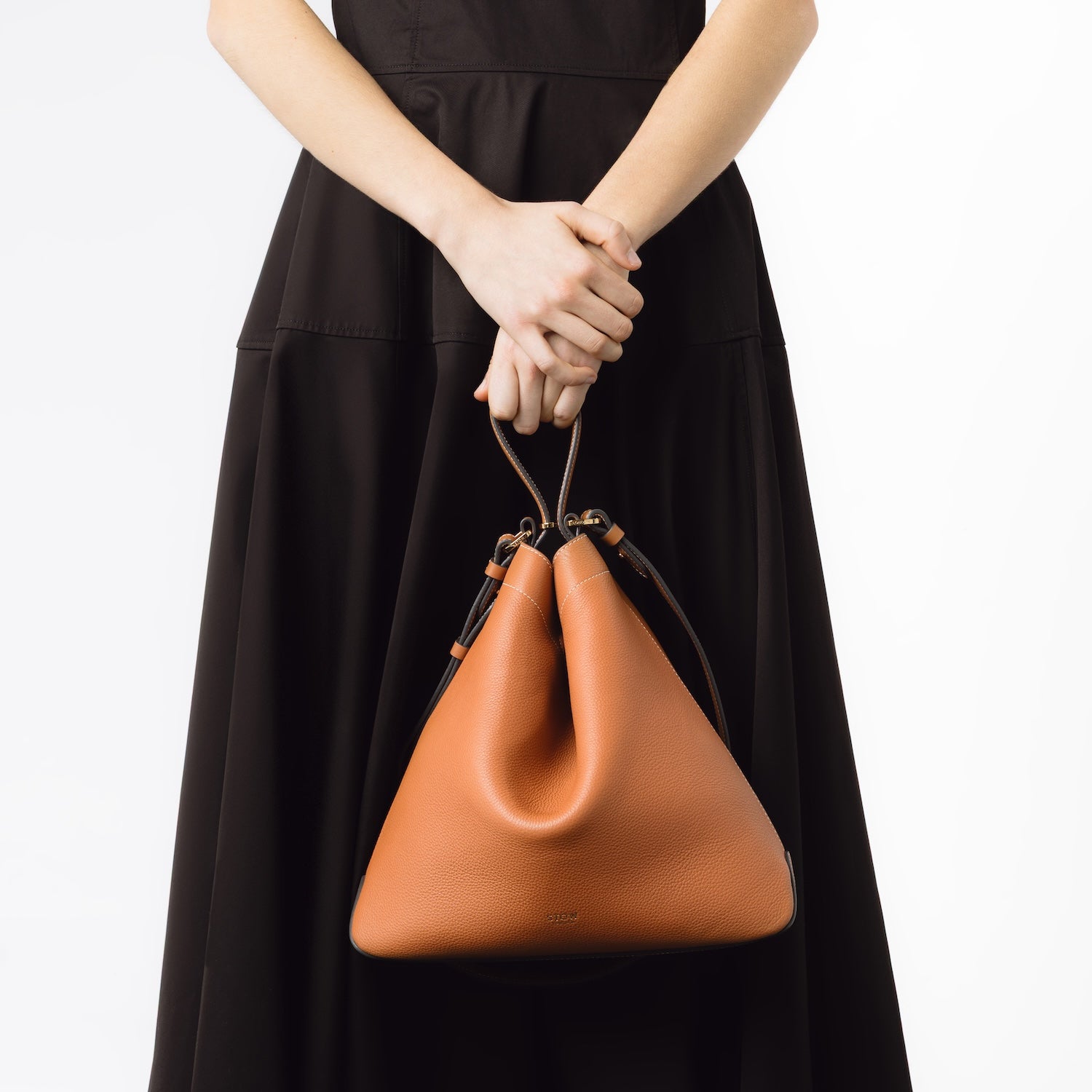 Model holding STOW London's Curve bag in Earth Tan by its top handle at her front featuring gold-coated stainless steel hardware on a white background