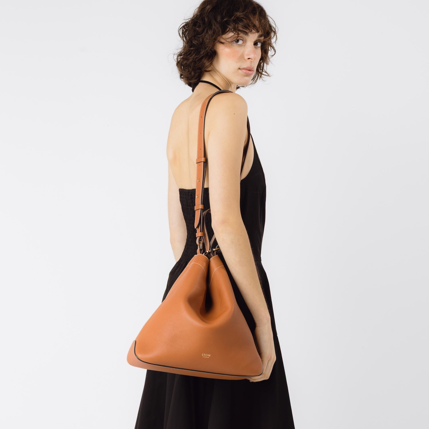 Model wearing STOW London's Curve bag in Earth Tan on her shoulder by its detachable strap featuring gold-coated stainless steel hardware on a white background